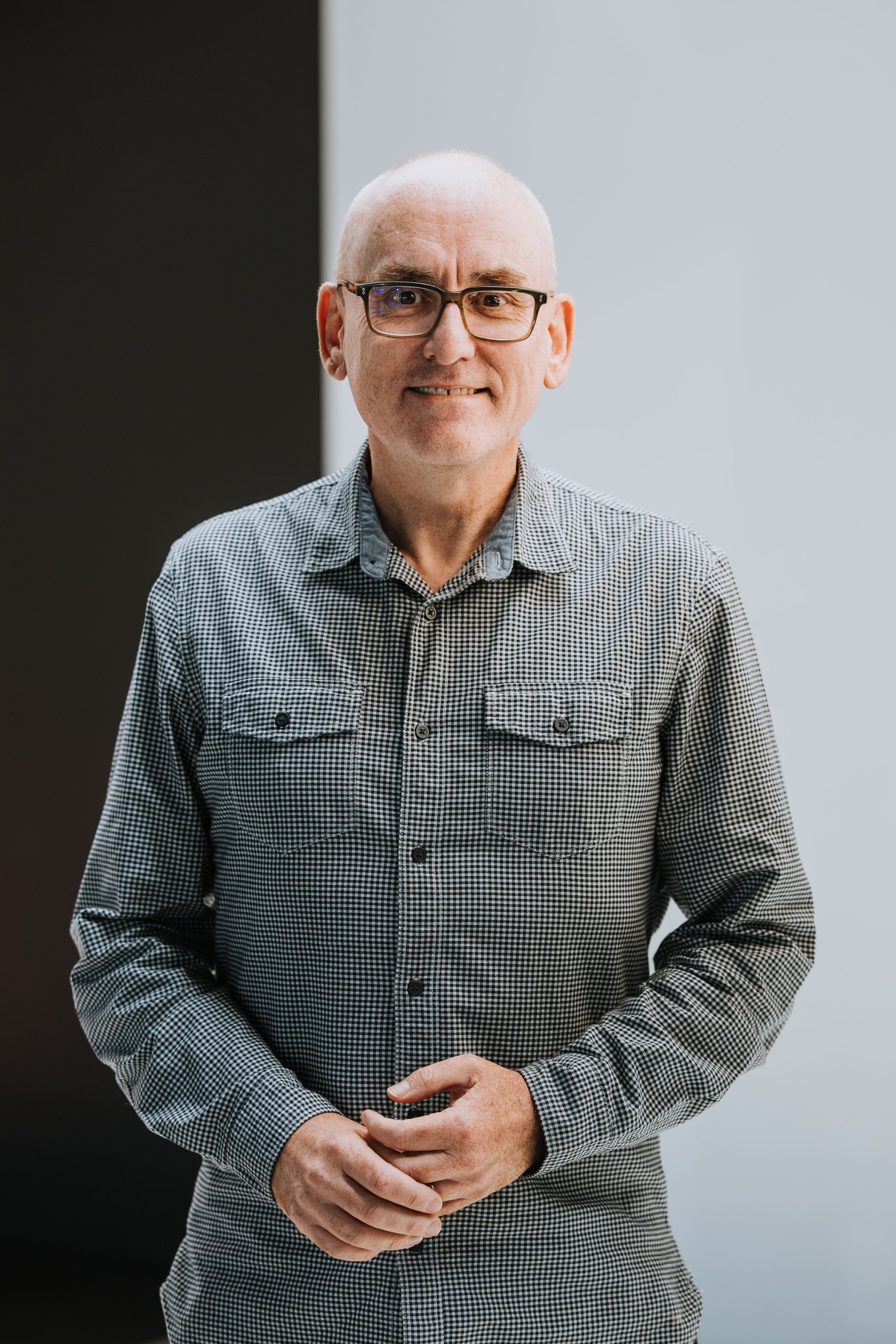 A middle-aged man with glasses and a bald head, wearing a black T-shirt and a black-and-white plaid shirt, smiling at the camera.
