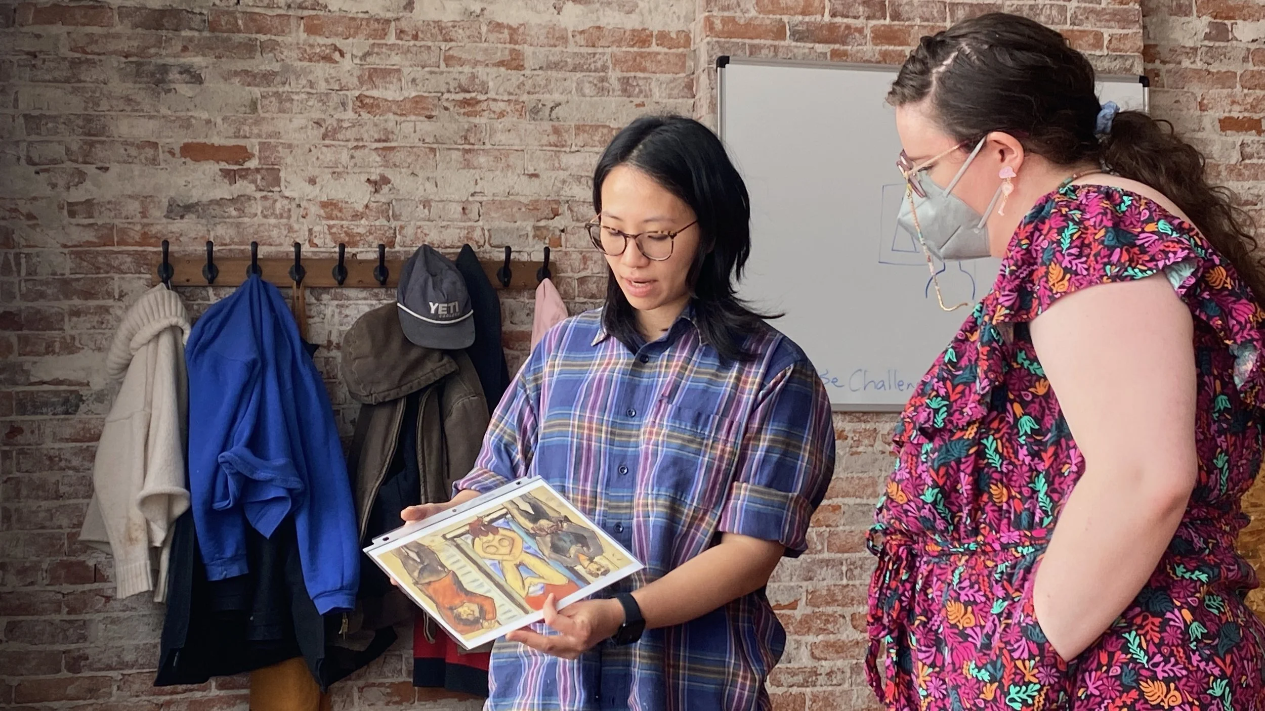 Two women stand in front of a brick wall, looking at a piece of artwork or a printed image. One woman is wearing glasses and a plaid shirt, holding the artwork, while the other woman is wearing a colorful floral dress and a face mask, observing. Behind them, there are hooks with coats and hats hanging on the wall, and a whiteboard in the background with some writing.