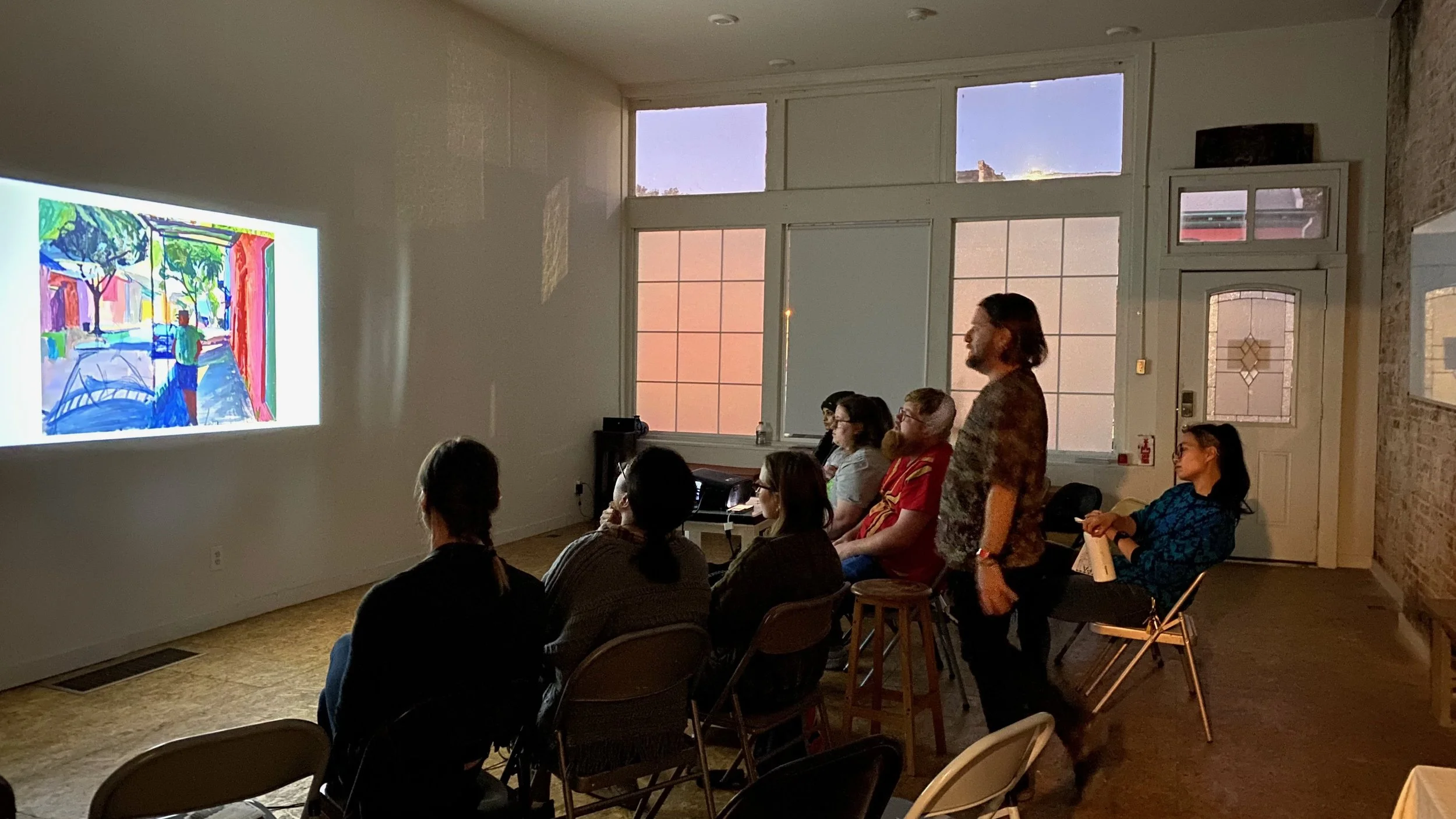A group of people seated and watching a colorful digital art presentation in a dimly lit room with large windows at sunset.