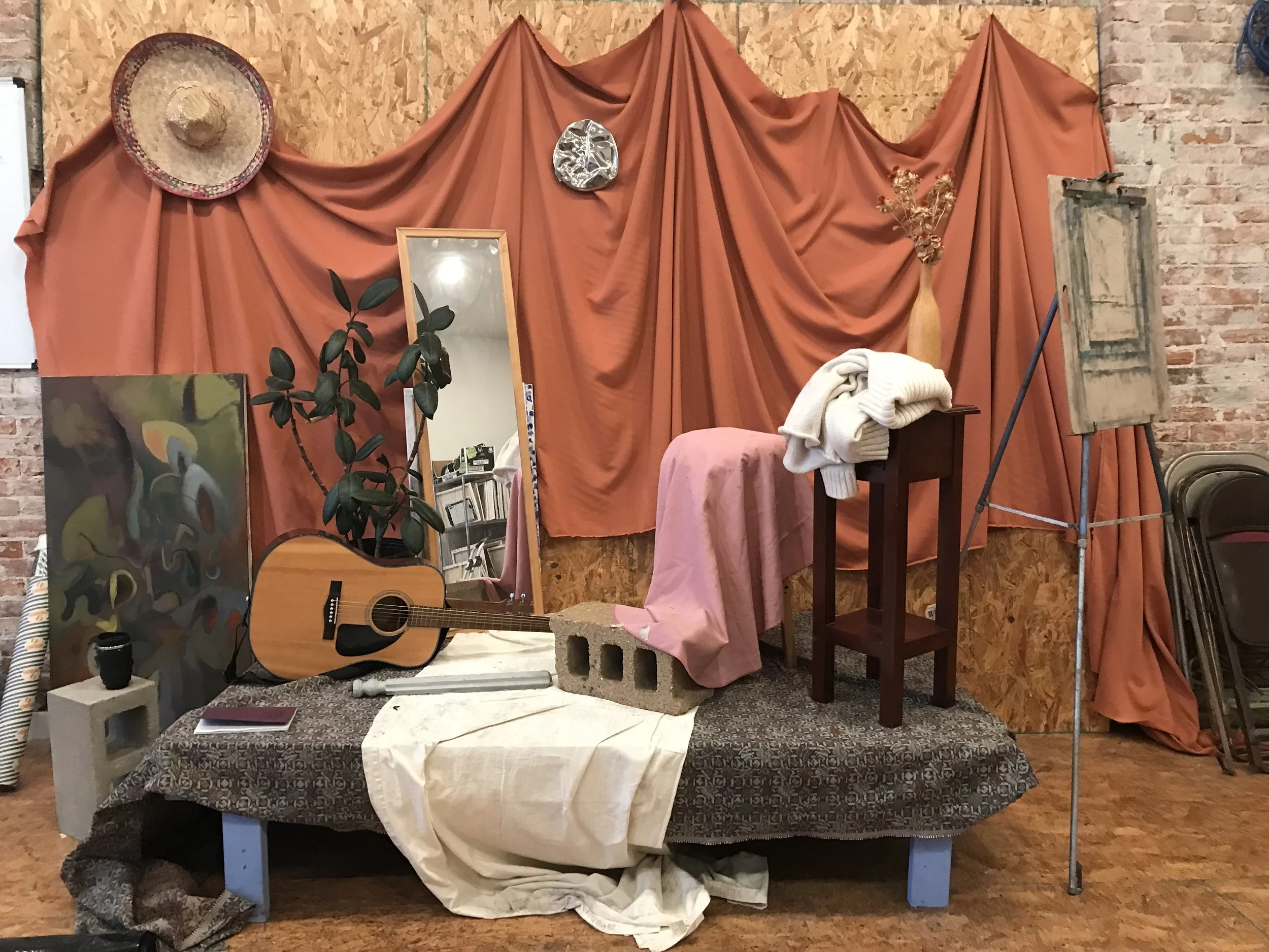 Eclectic studio setup with draped fabric, a mirror, a guitar, a plant, a painting, and various decorative objects on a platform.