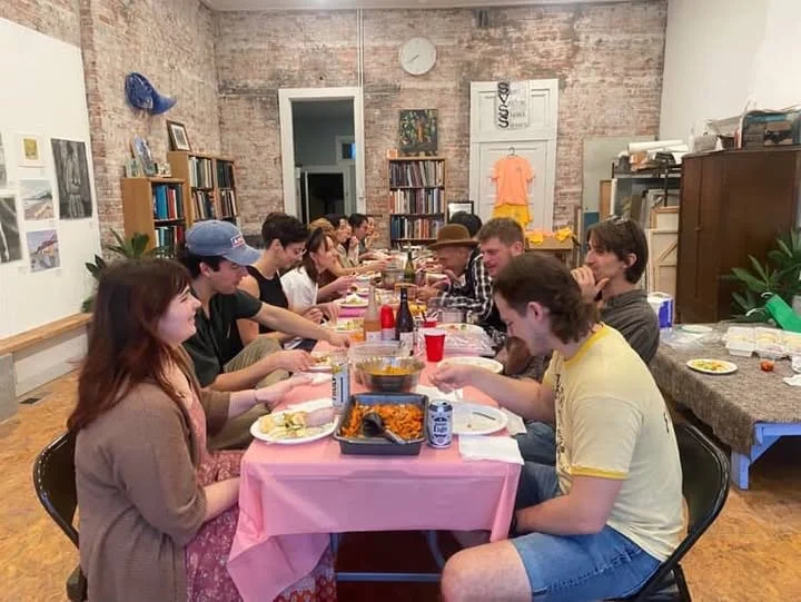 People sitting around a long dining table enjoying a meal in a casual, cozy room with brick walls and bookshelves.