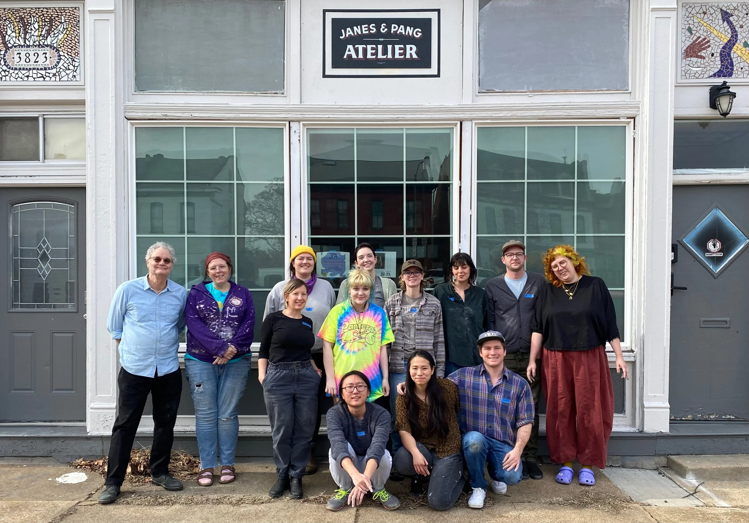 Group of 14 people standing and kneeling in front of a building with a sign that reads 'Janes & Pang Atelier'.