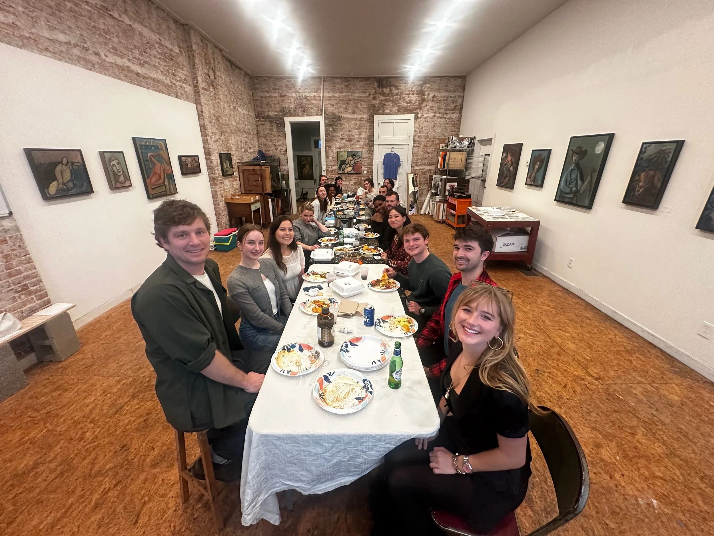 People gathered around a long dining table enjoying a meal in an art gallery or studio with artworks on the walls.