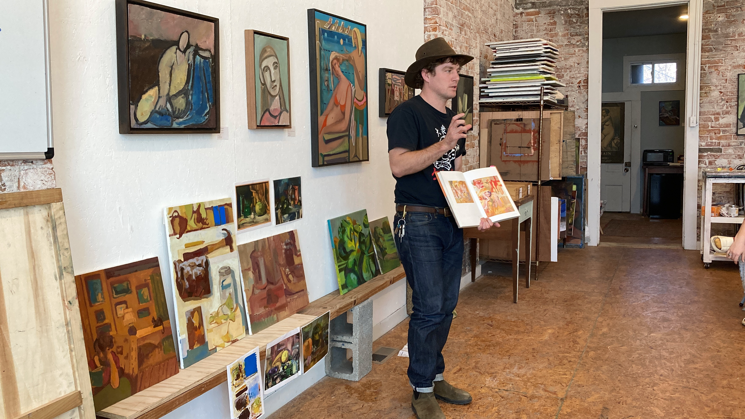 A person in a brown hat and casual clothes presenting to an audience inside an art gallery. They are holding an open book and gesturing with their right hand. Various paintings are displayed on the walls and on a shelf, with a partially visible doorway and window in the background.