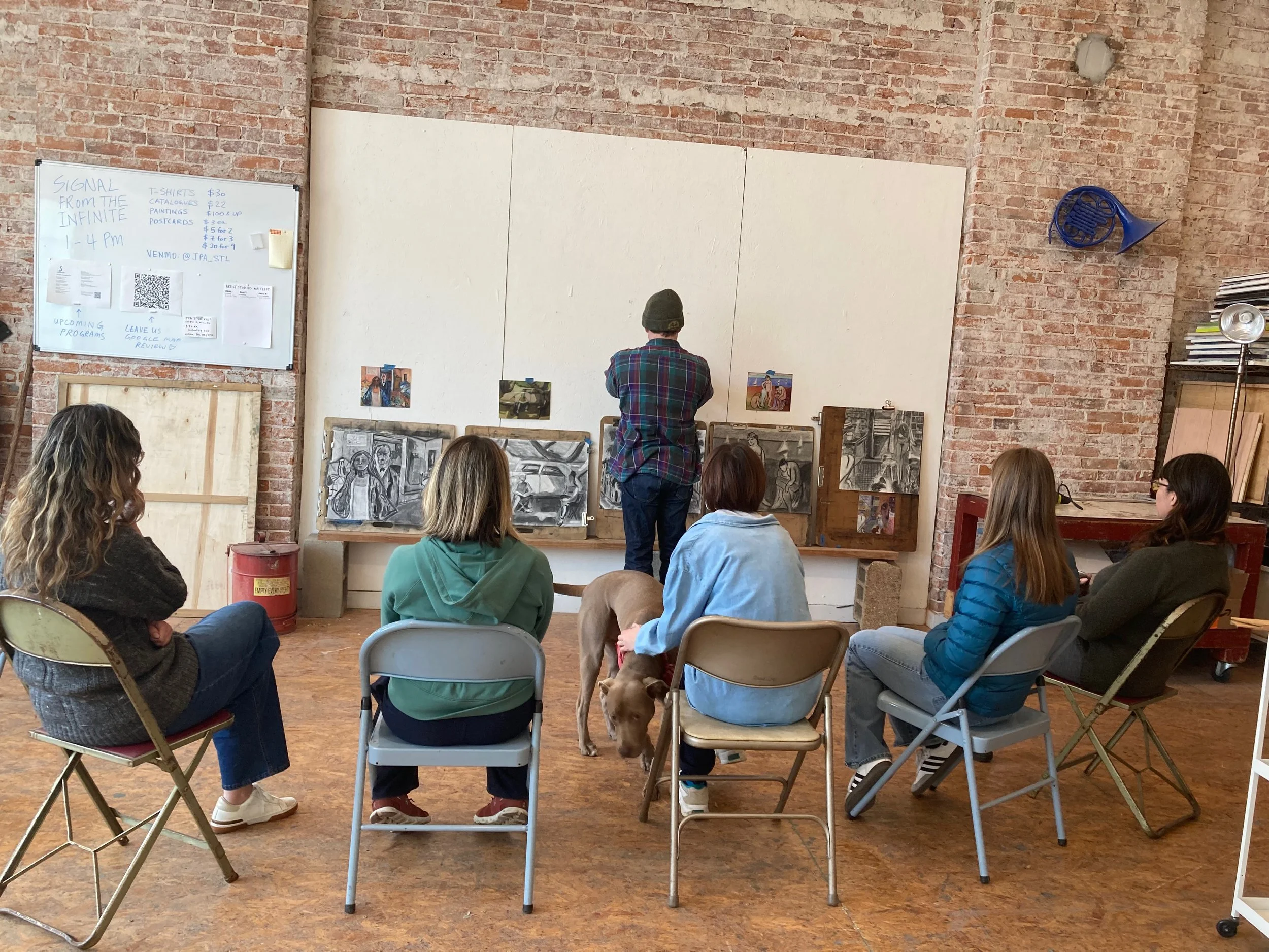An art class with six women seated on chairs watching a man in a plaid shirt and beanie draw sketches on large white paper on a wall. A dog is standing near one of the women.