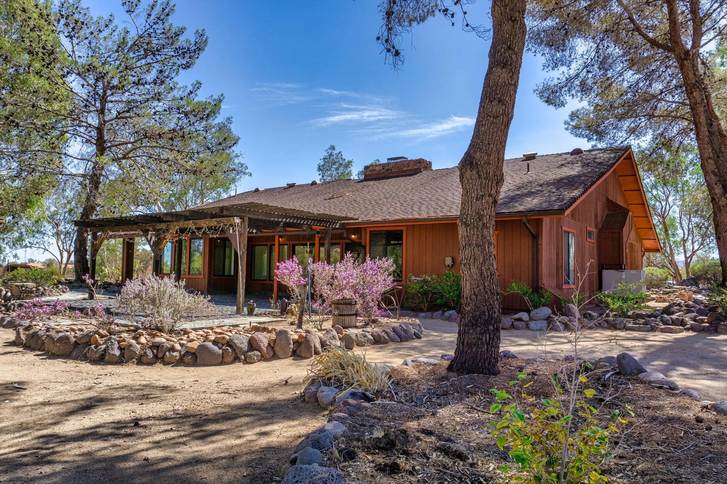 A wooden house with a shaded porch, surrounded by trees, bushes with pink and purple flowers, and a rocky landscape under a blue sky.