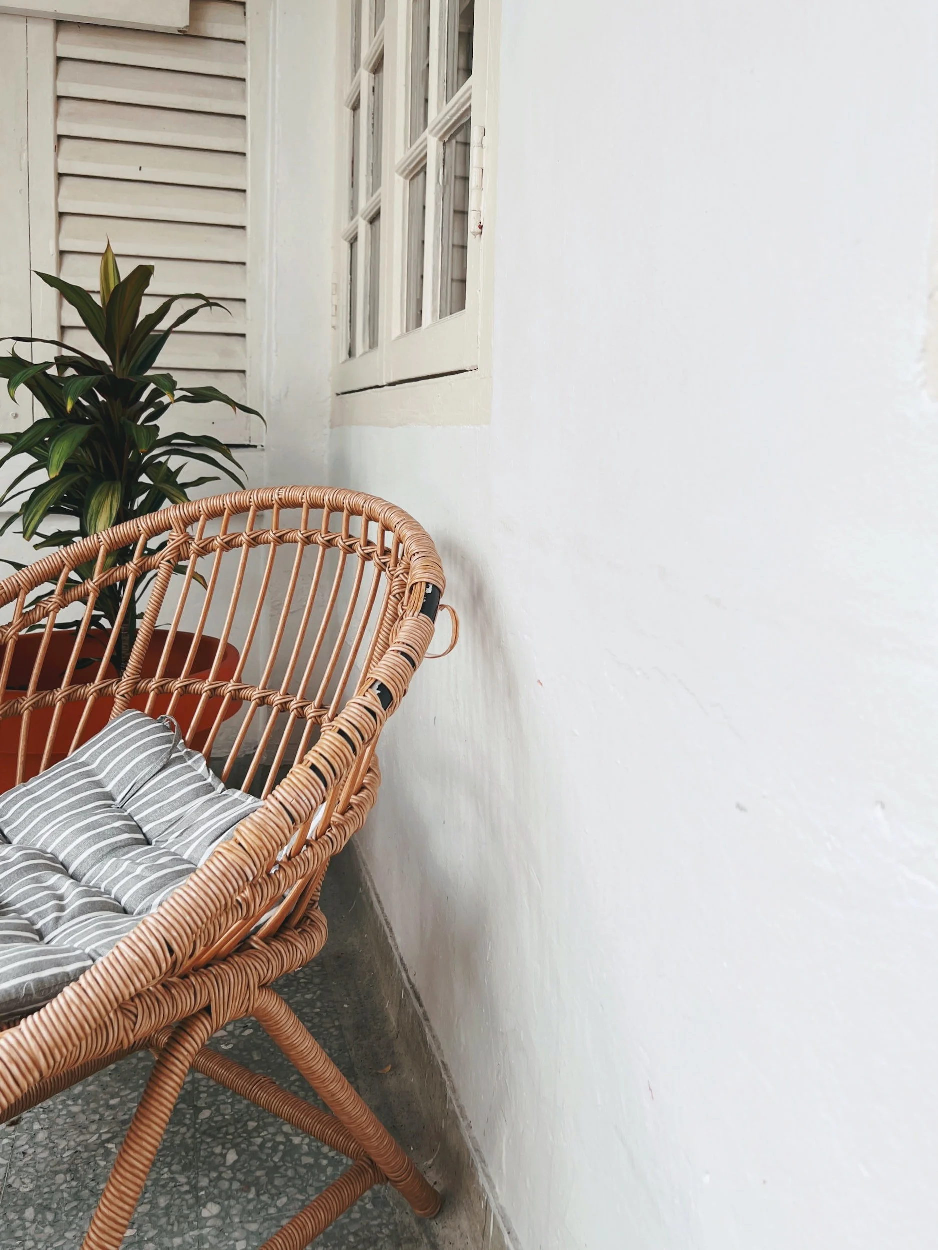 Wicker chair with gray and white striped cushion next to a potted plant on a porch with white walls and window.