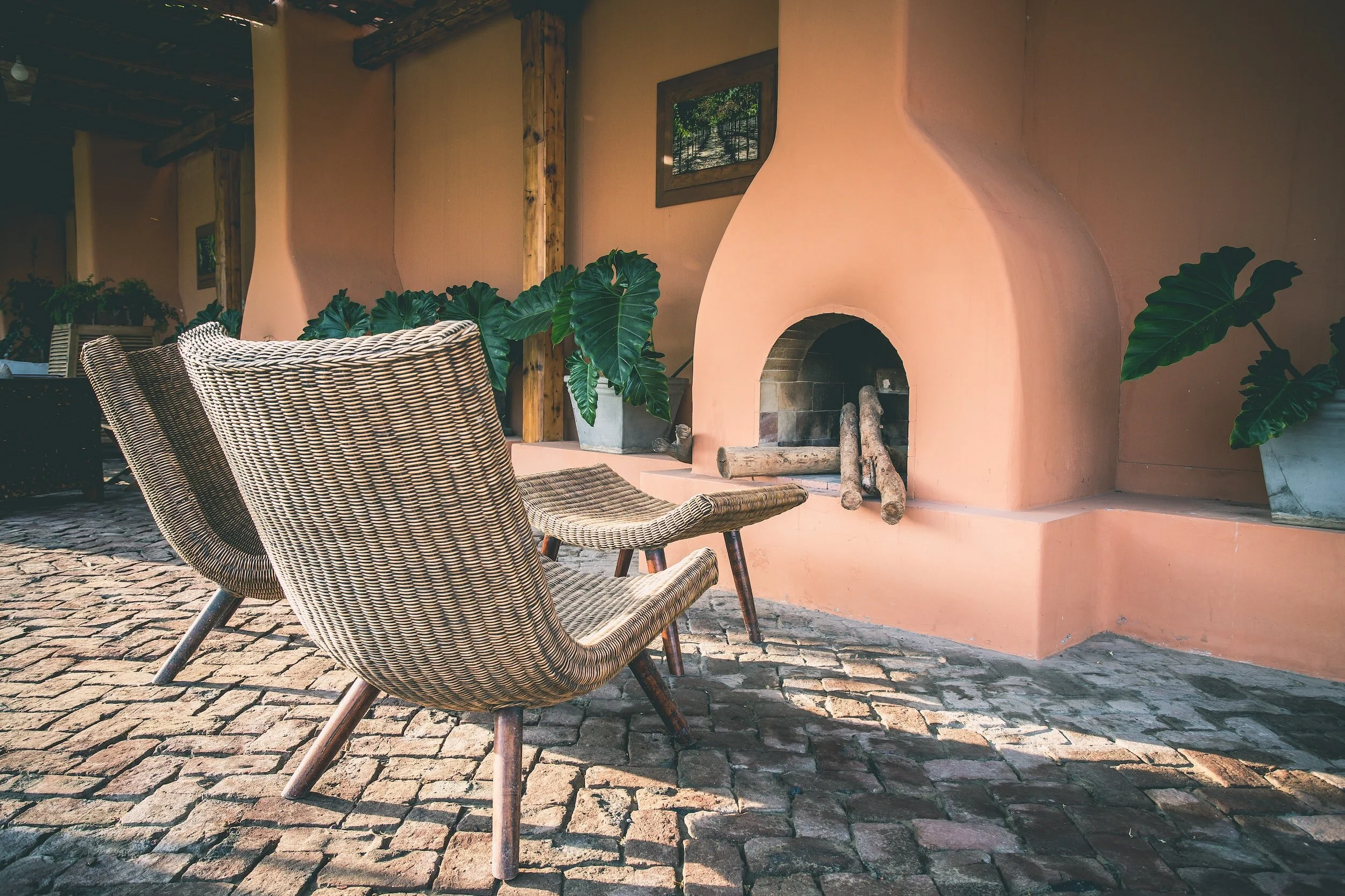 Two wicker chairs and a small wooden table in front of a pink stucco fireplace with logs. Potted plants and a framed picture on the wall in a rustic outdoor setting.