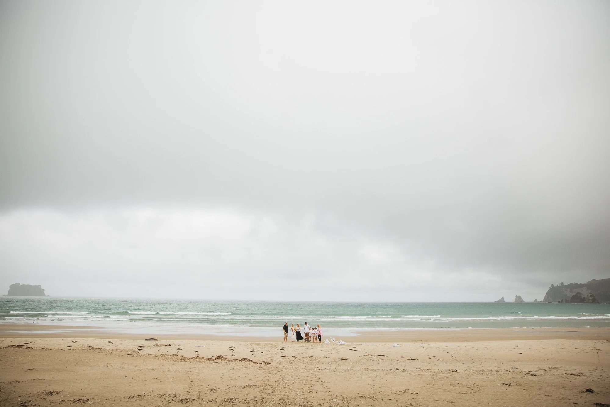 Family gathered on Whangamatā Beach during Ross and Renee’s stormy vow renewal, holding clear umbrellas in the rain.