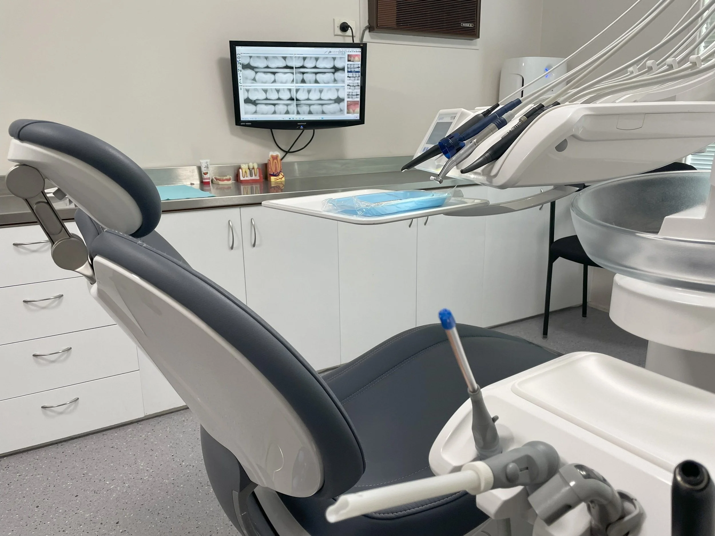 Dental exam room with an empty dental chair, dental tools, a monitor displaying dental X-rays, and medical supplies.