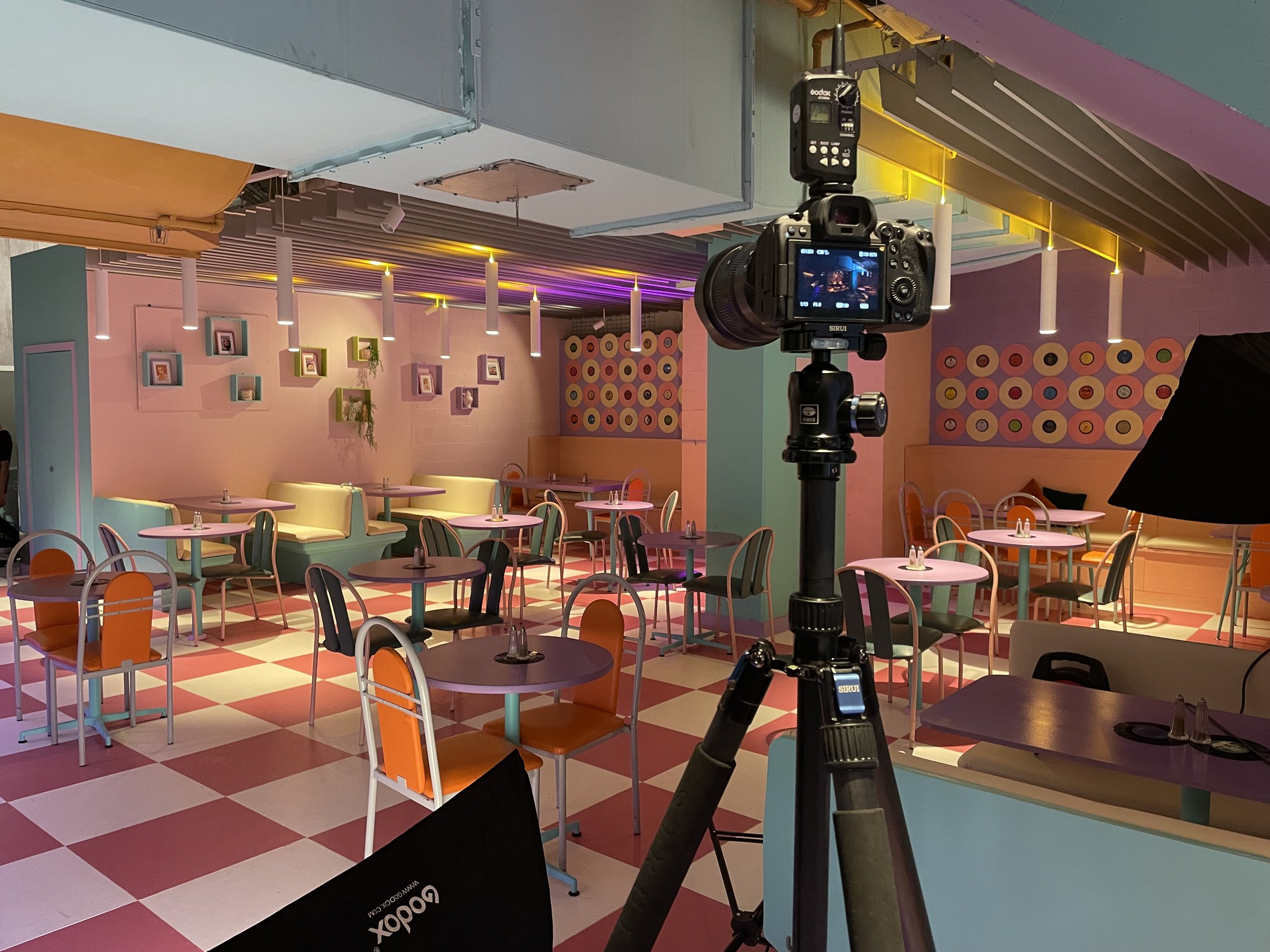 Interior of a colorful, retro-style cafe or restaurant set up for photography, with a camera on a tripod, pastel chairs and tables, checkered floor, and decorative wall art. Large softbox light is also visible.