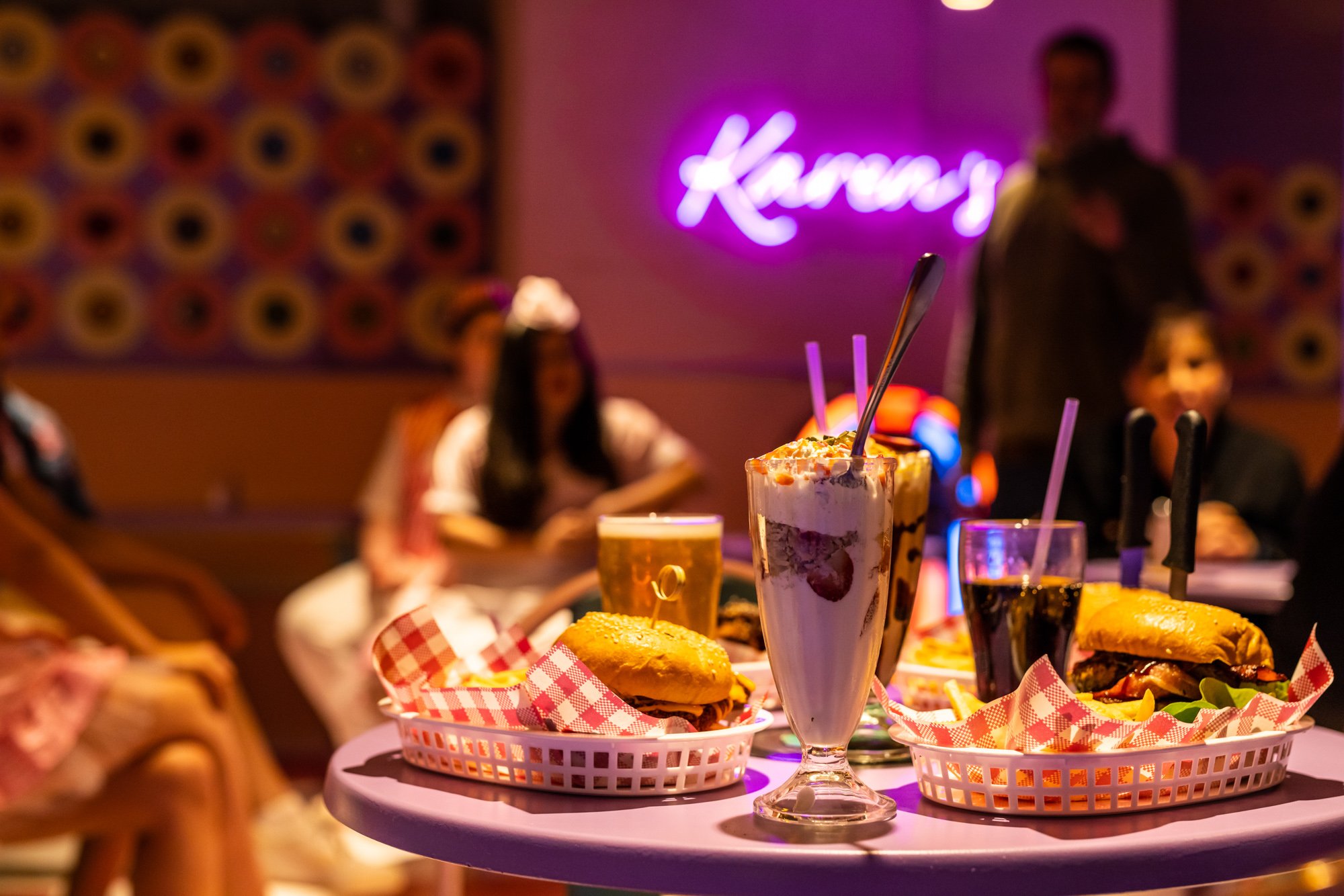 A table with various fast food items including sandwiches and milkshakes in a neon-lit restaurant with people in the background.