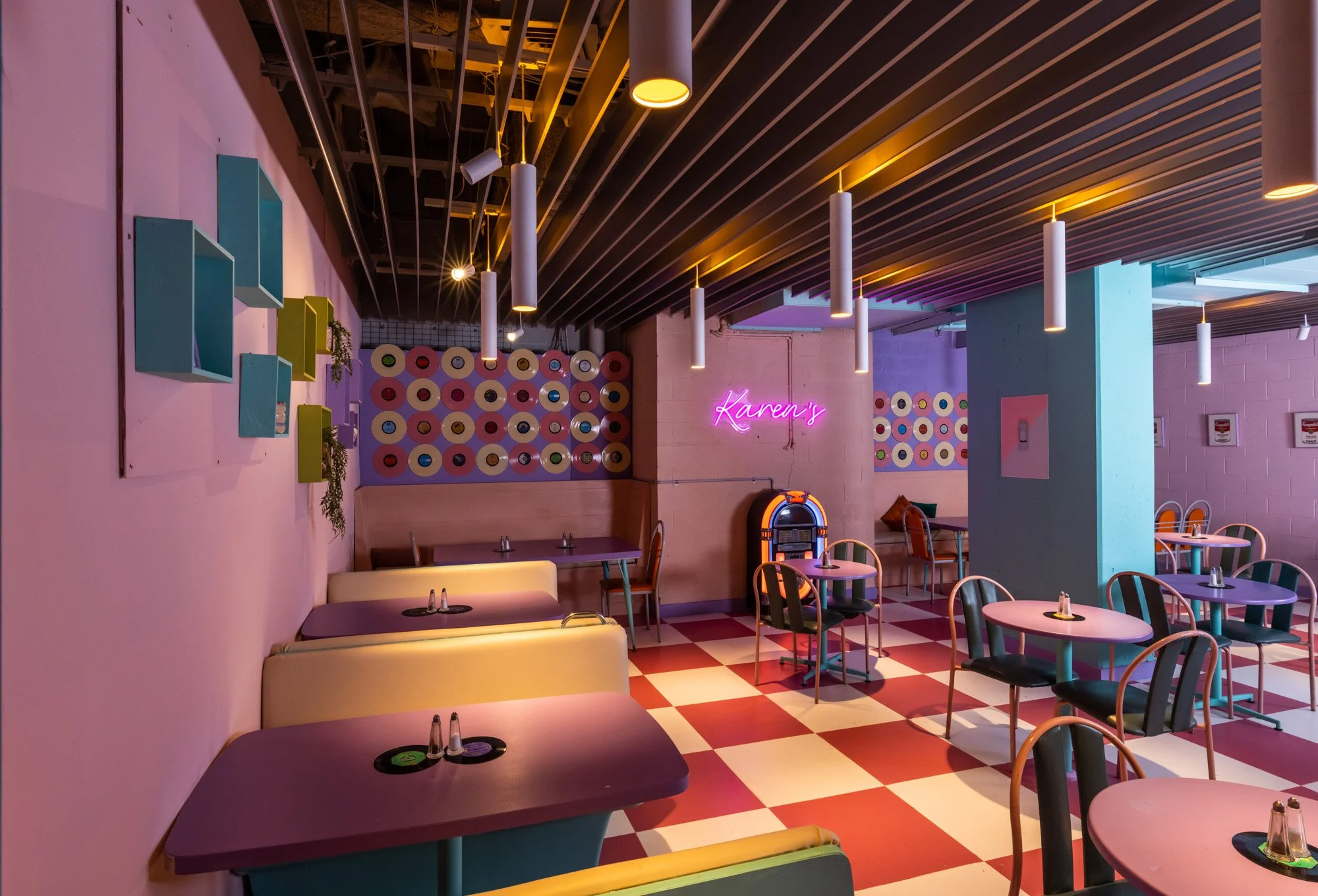 Colorful retro-style diner with pink and blue walls, checkered red and white floor, round tables with chairs, neon sign that reads "Karen's", jukebox, and hanging white pendant lights.