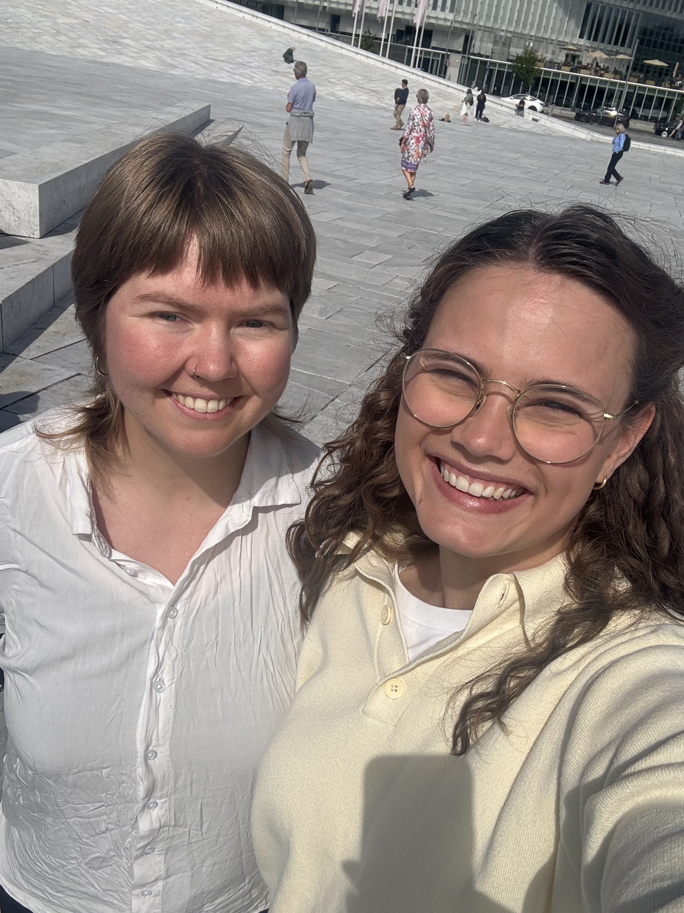 Øyunn S. Kåset er til venstre i bildet og Tora Tveiten til høyre. Begge smiler bredt og myser mot solen som skinner. I bakgrunnen ser man marmorsteinene på Operaen i Oslo