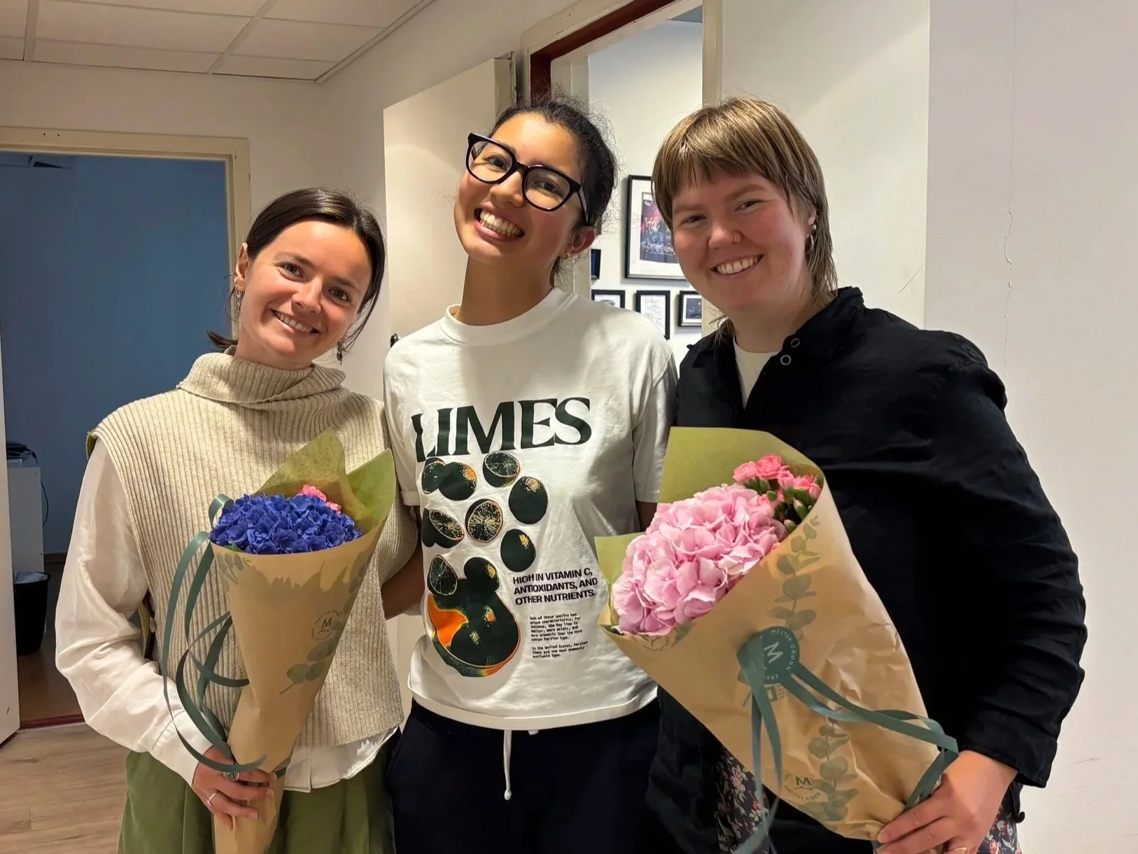 Ida Reffhaug står til venstre i bildet og holder en bukett med blå blomster. I midten står Victoria Wang med armene rundt Ida på sin venstre side og Øyunn på sin høyre side. Øyunn holder en bukett med rosa blomster. I bakgrunnen ser man kontoret.