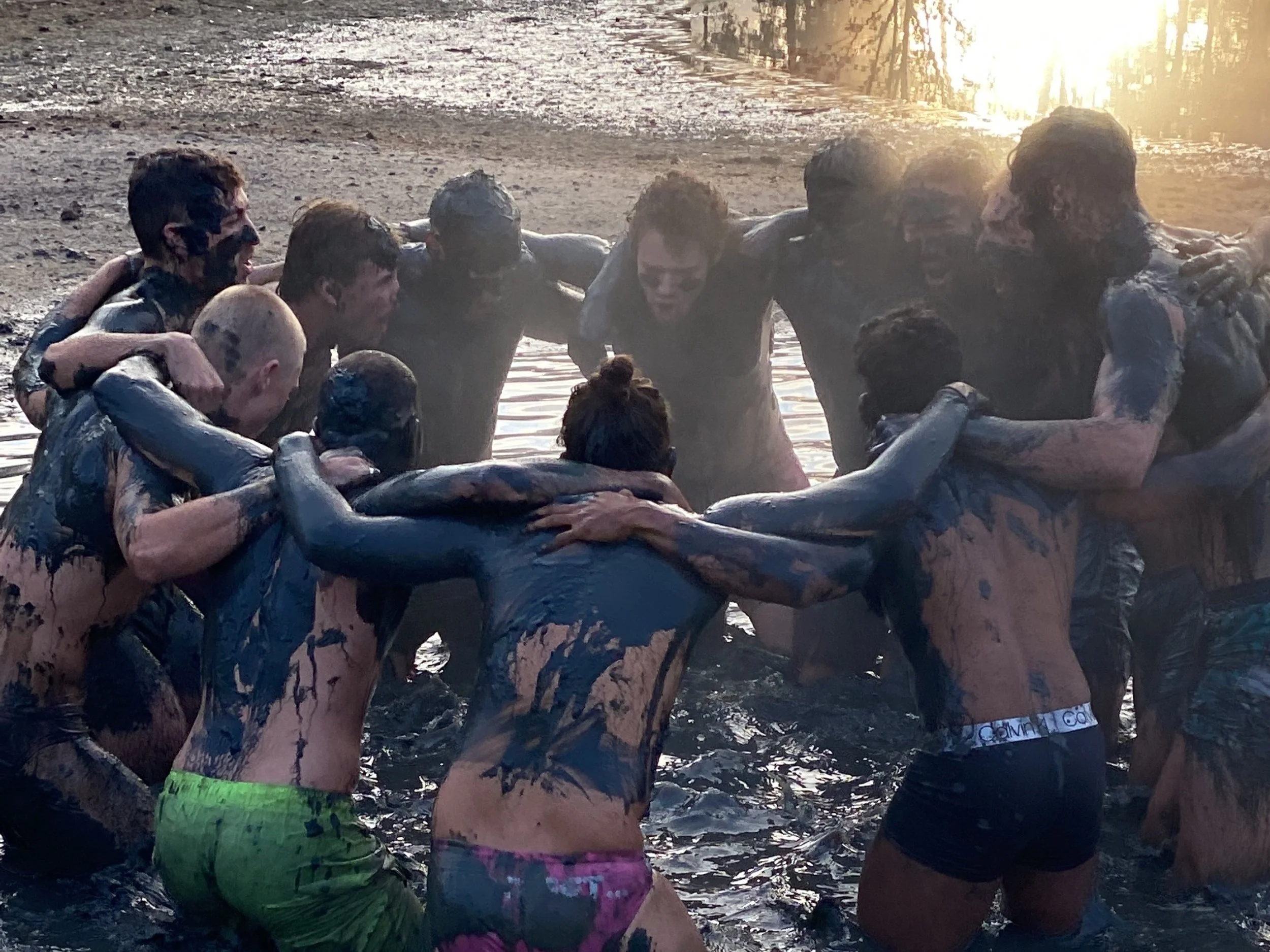 A group of men covered in mud with arms around eachother, surrounded by nature