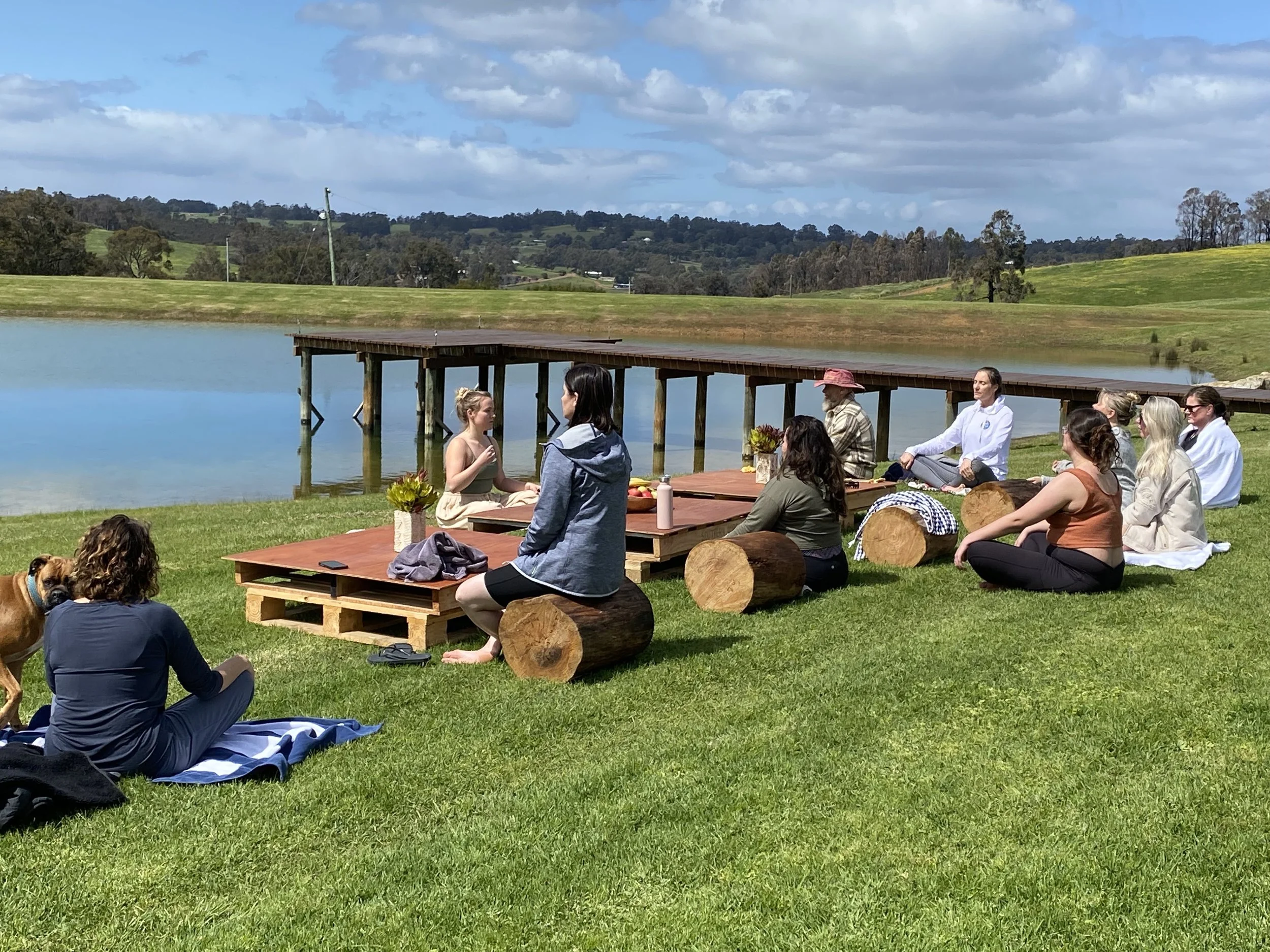 a group of women, sitting on wooden stumps, outdoors in nature by a dam, connecting with eachother