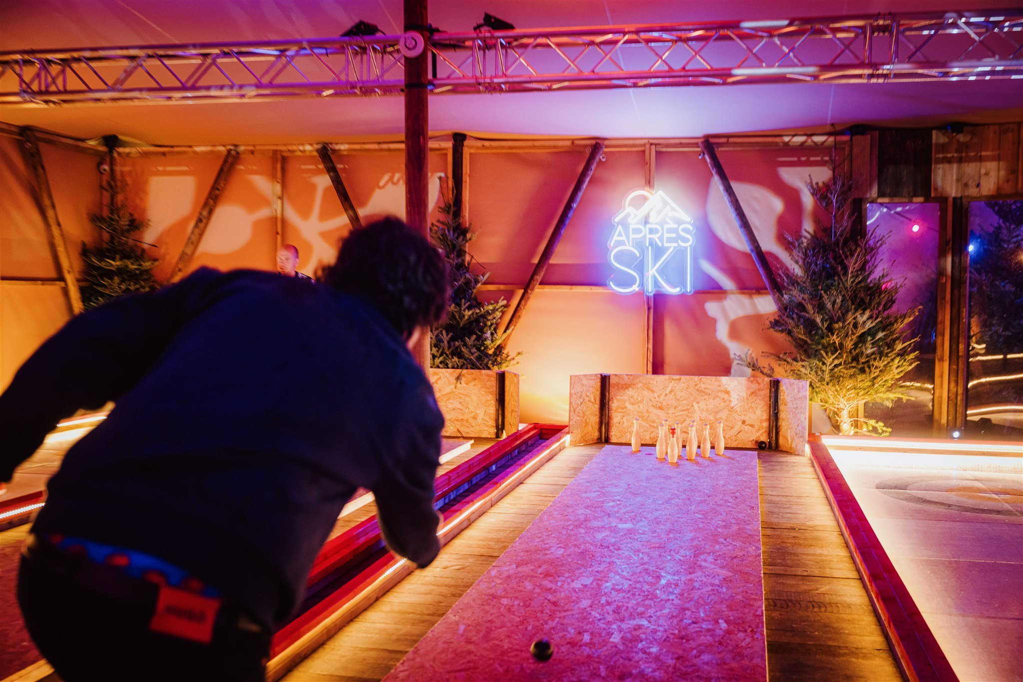 Person aiming to bowl at a neon-lit alley with a sign reading 'Après Ski' and decorated with Christmas trees.