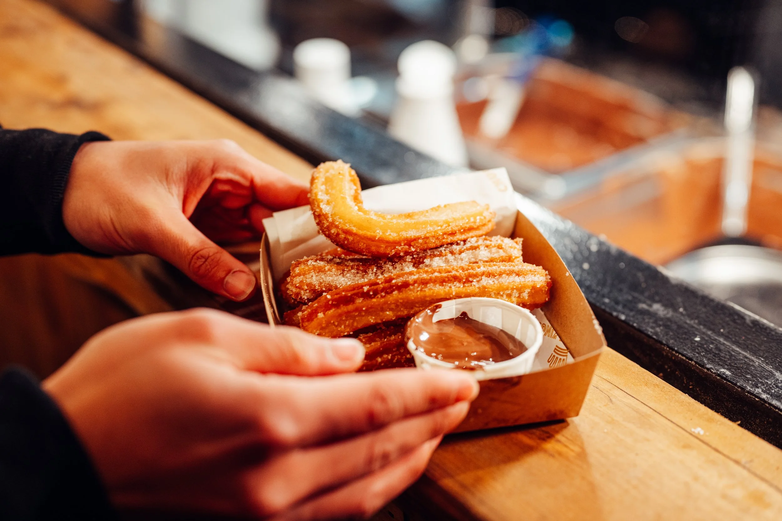 Person holding a cardboard tray of fried churros with a cup of chocolate dipping sauce.