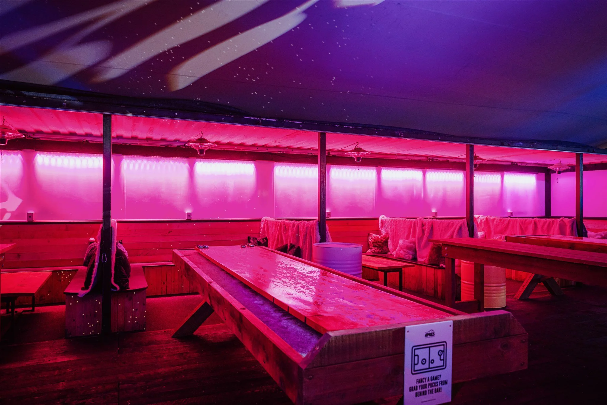 Indoor recreational area with pink lighting, wooden benches, and a shuffleboard court.