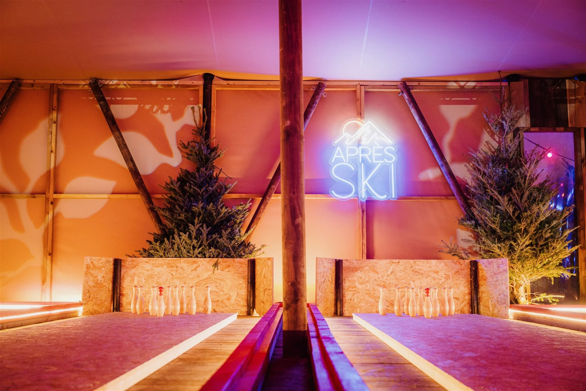 Indoor scene of a ski-themed bowling alley with two lanes, decorated with pine trees and a neon sign that reads 'Après Ski'. The lighting has an orange and purple hue.