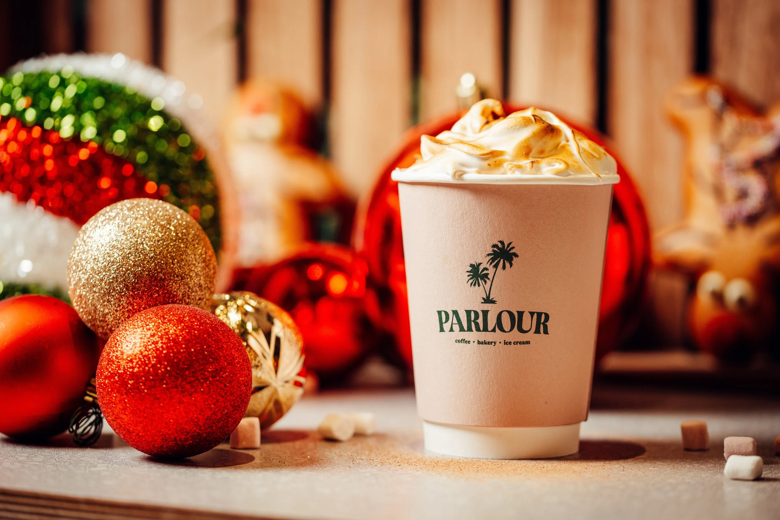 A cup of whipped cream topped with caramel sauce from Parlour, surrounded by Christmas ornaments including red, gold, and glittery baubles, with Christmas decorations and a wooden backdrop in the background.