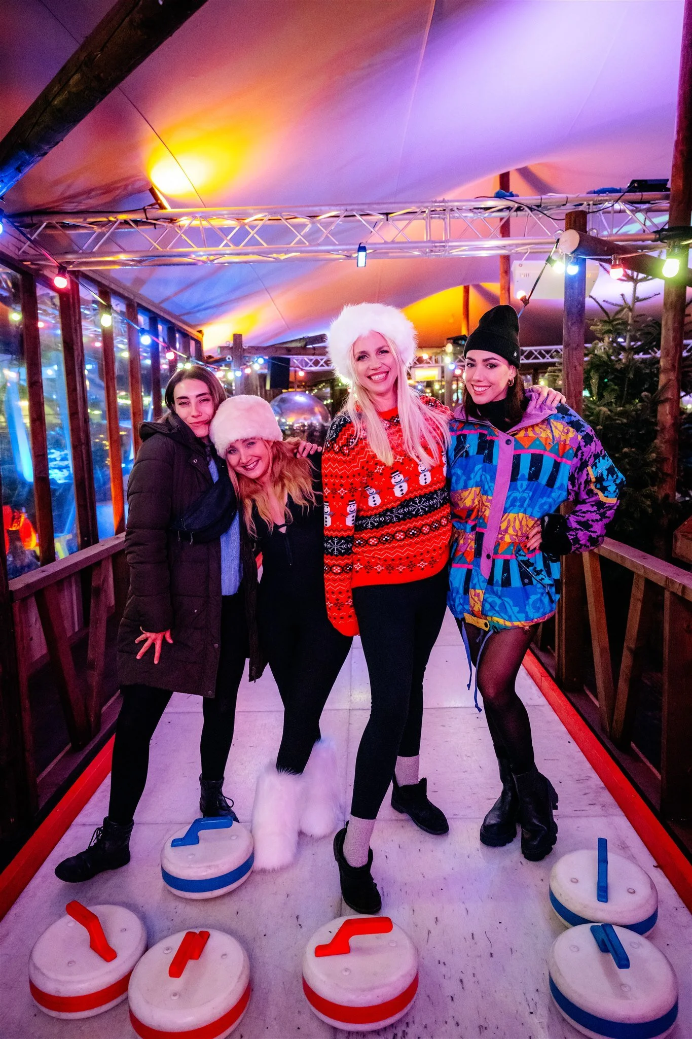 Four women posing together on an ice rink decorated with colorful string lights, wearing winter clothes and festive sweaters. One woman is wearing a Santa hat, and a dog with a fluffy white tail is in front, playing hockey.
