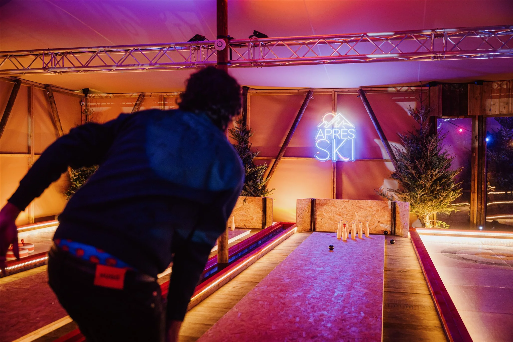 A person playing a game of bowling or skittles at an indoor arcade venue with a neon sign that reads 'APRES SKI'. There are small decorative trees and the scene is lit with colorful ambient lighting.