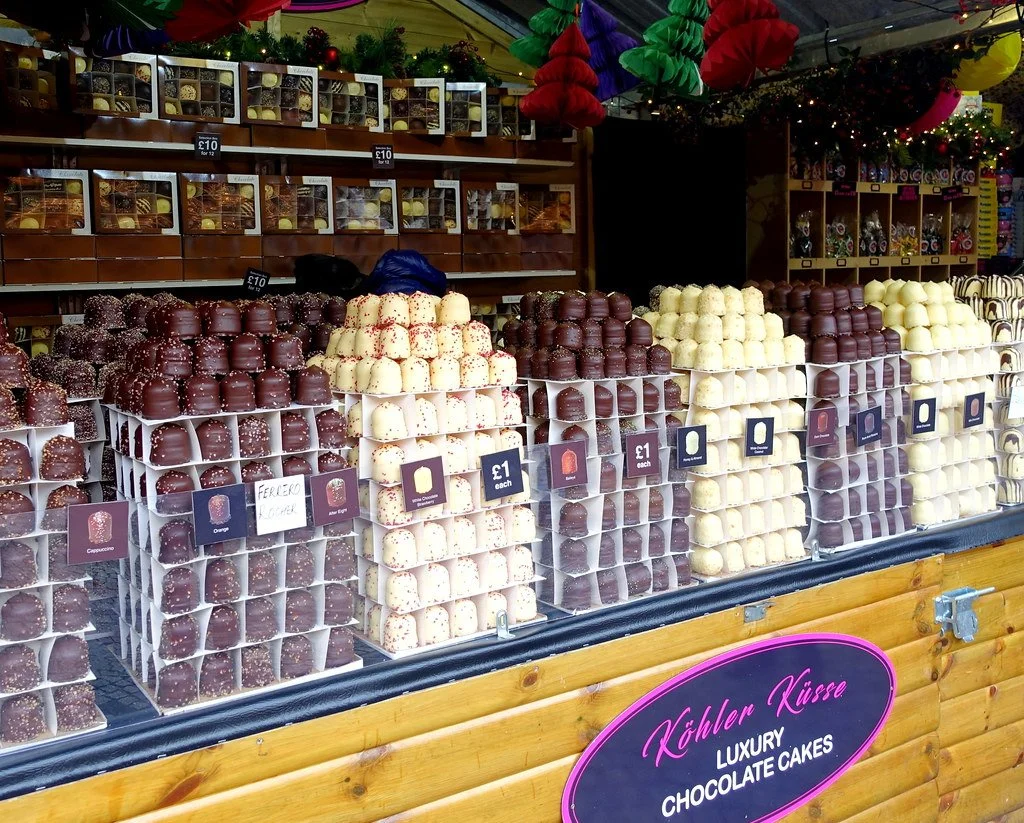 Display of luxury chocolate cakes arranged in front of a wooden stall at a market, with colorful decorations overhead and signs indicating prices.