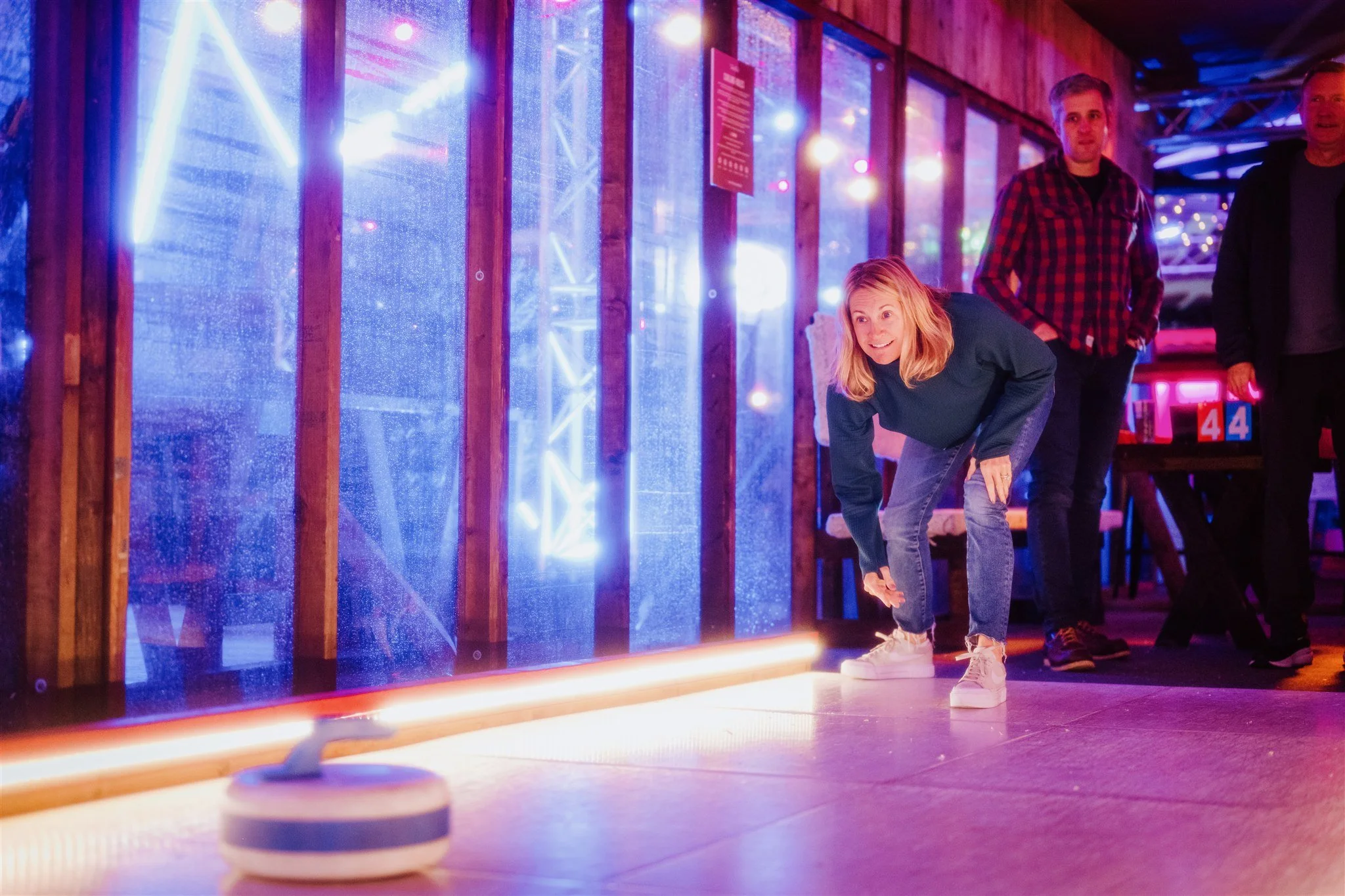 A woman playing curling in a rink with colorful lighting, with two men watching nearby.