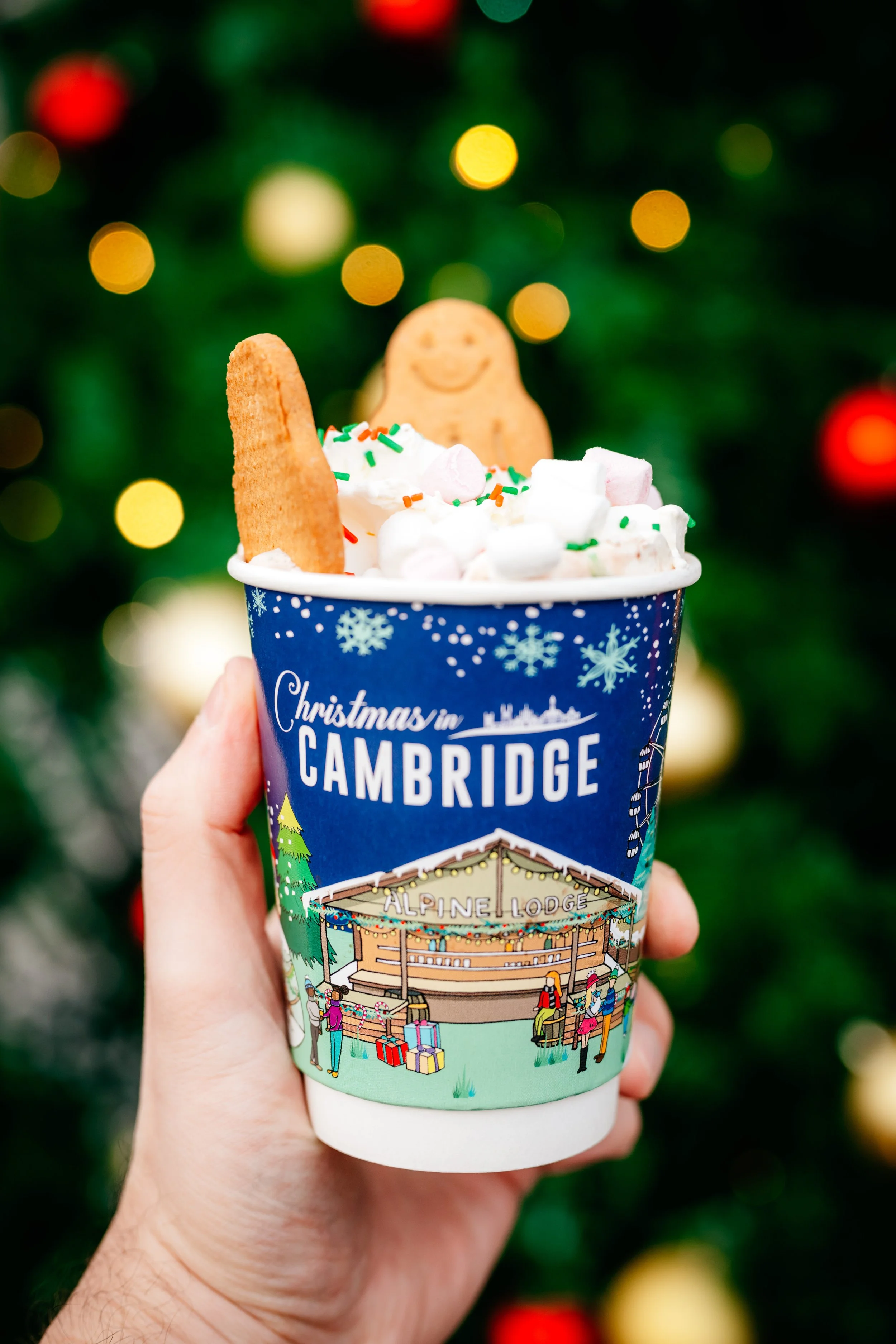 A hand holding a holiday-themed hot chocolate cup topped with whipped cream, marshmallows, a cookie, and a smiling gingerbread cookie in front of a decorated Christmas tree with red and yellow lights.