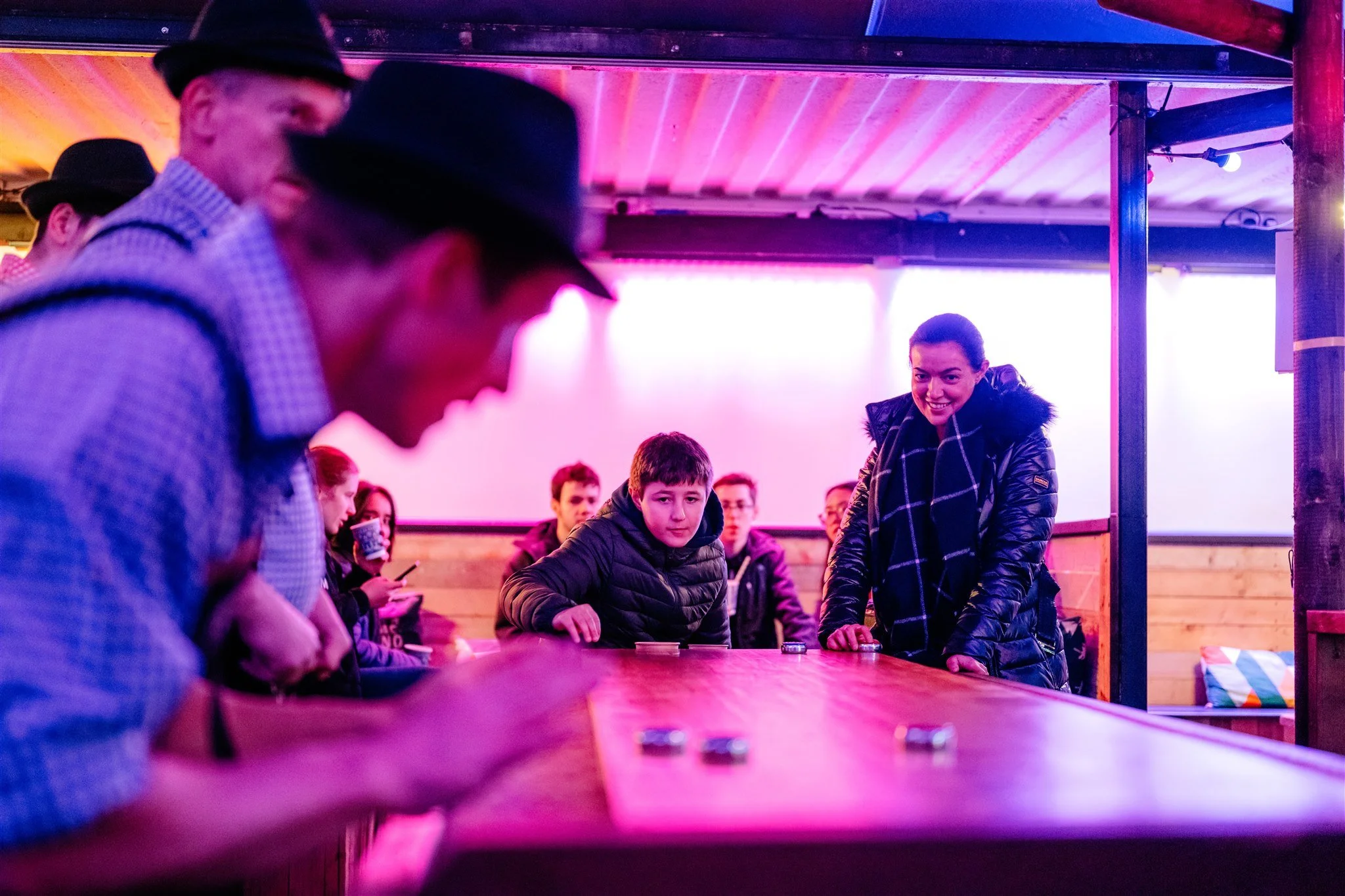 A group of people, including kids and adults, gathered around a long wooden table in a room with colorful lighting. Some individuals are engaged in a game or activity, with a woman smiling and leaning over the table, and a boy looking directly at the camera. The background shows a bright screen and wooden walls.