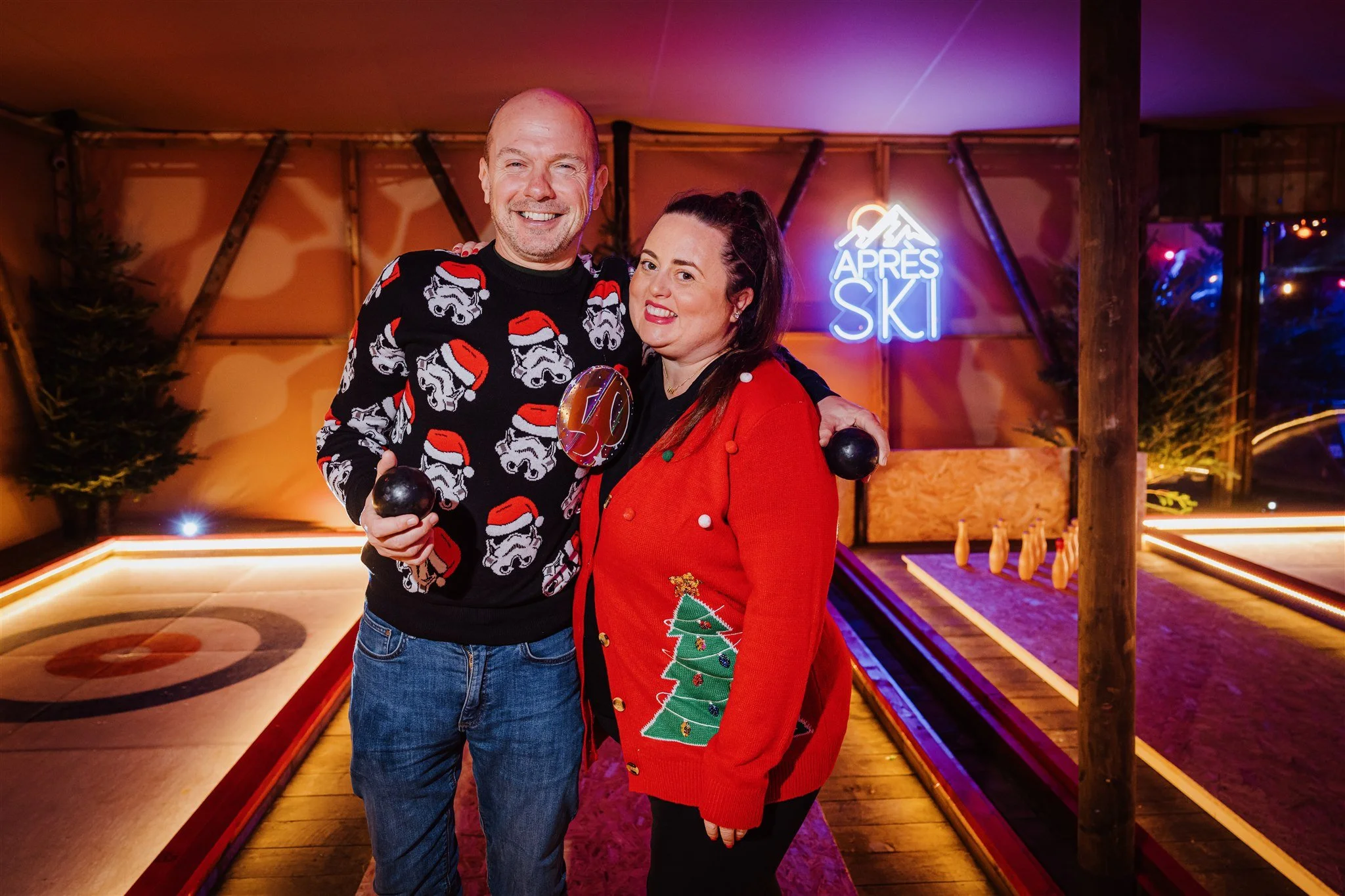 A man and woman are smiling and holding black bowling balls in a decorated indoor bowling alley with a Christmas theme. The man is wearing a sweater with stormtroopers wearing Santa hats, and the woman is wearing a red cardigan with a Christmas tree 