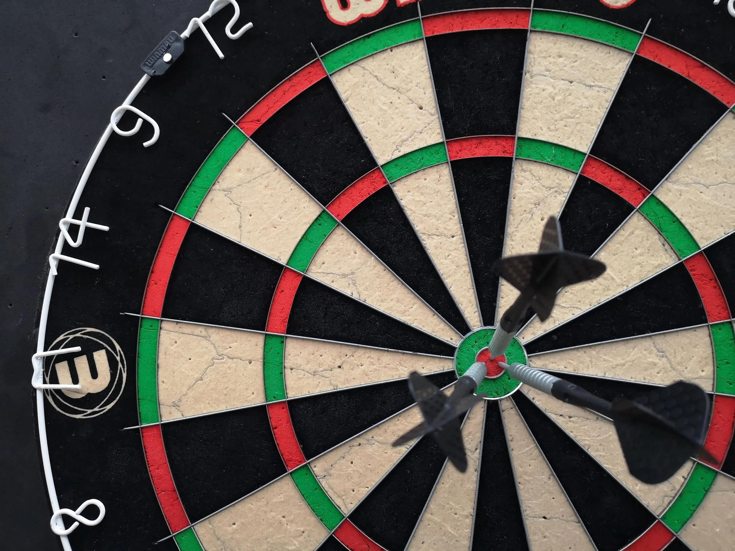 Close-up of a dart hitting the bullseye on a dartboard with red, green, black, and beige sections, with a white wire surrounding the dartboard's numbers.