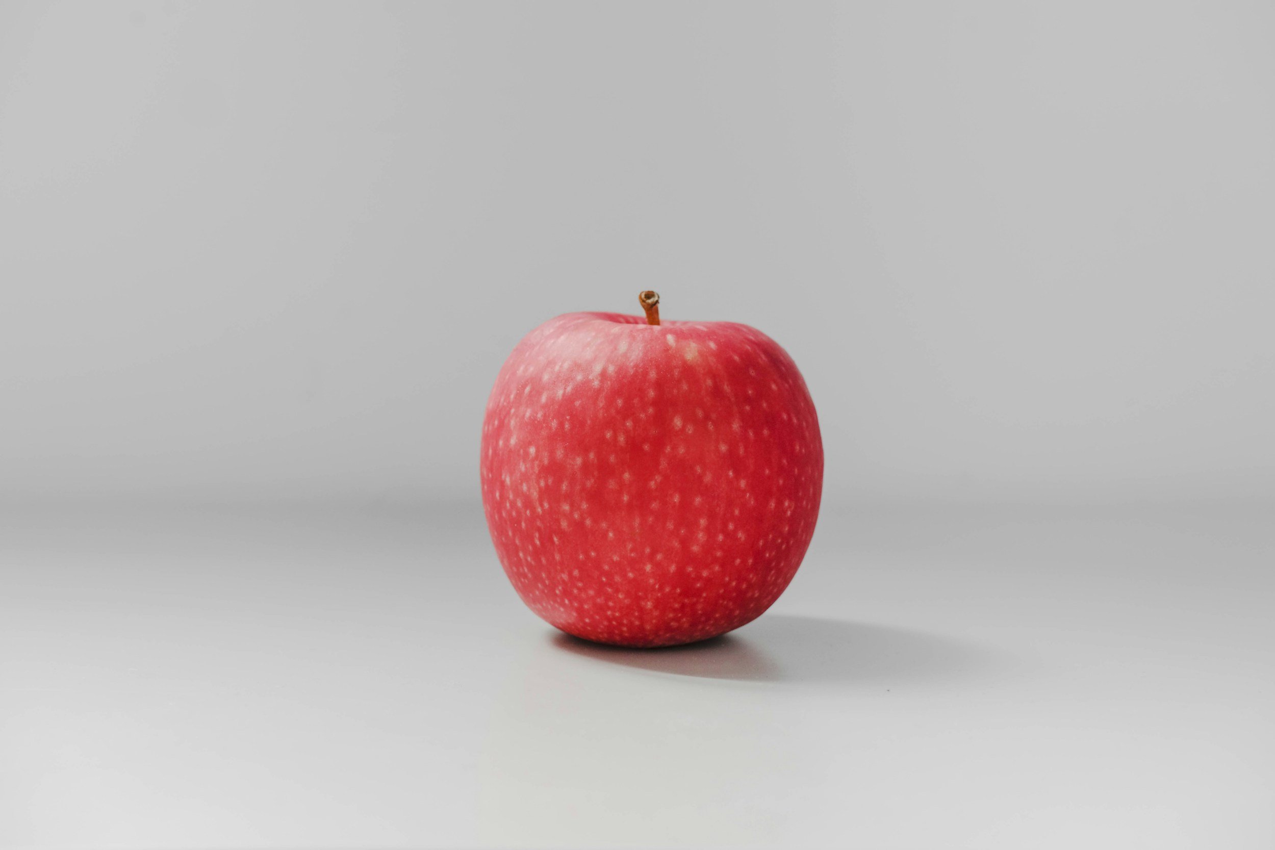 Red apple with a small brown stem on a plain gray surface with a gray background.