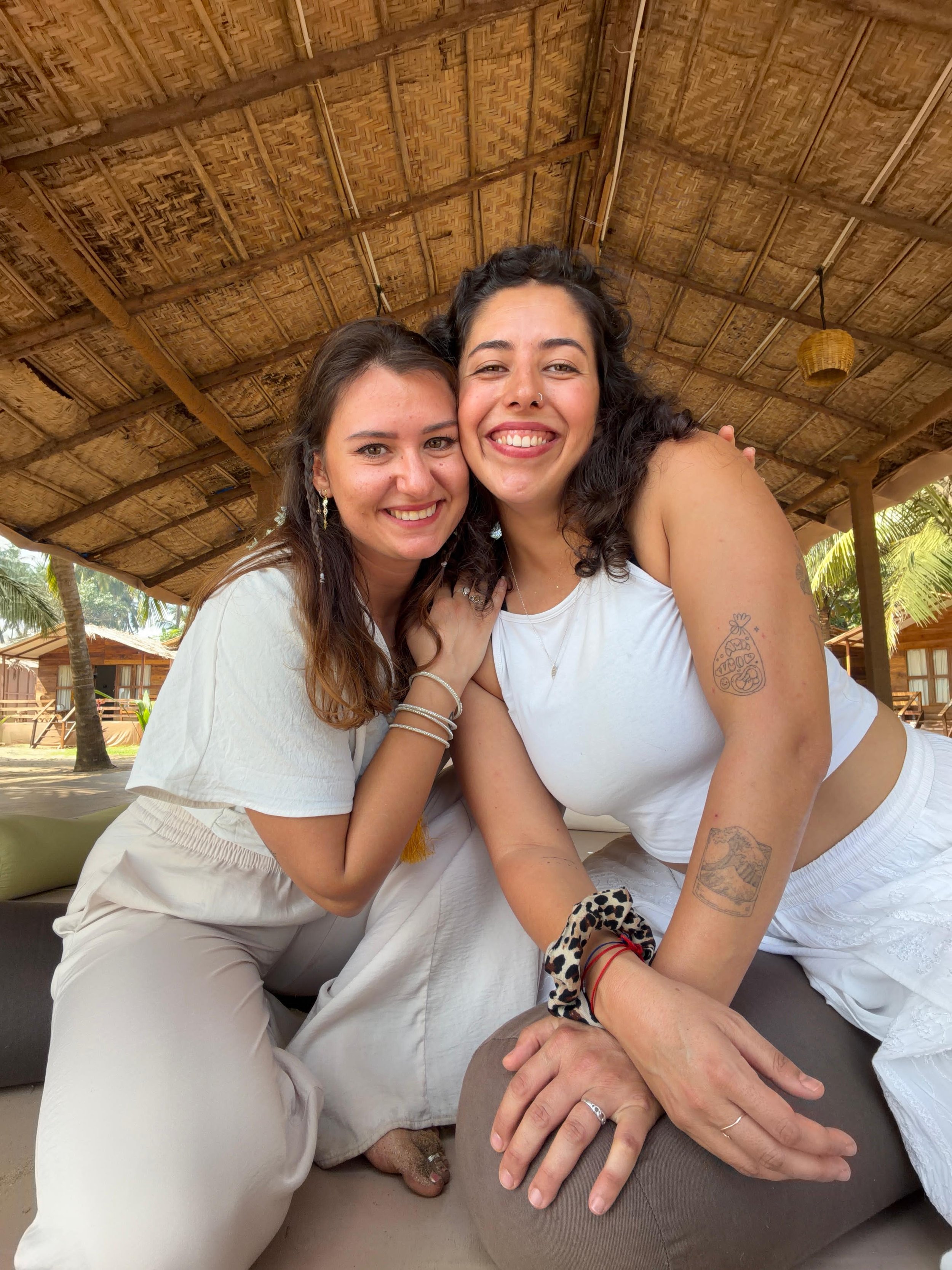 Two women sitting close together, smiling, under a thatched roof, with tropical palm trees and wooden huts in the background.