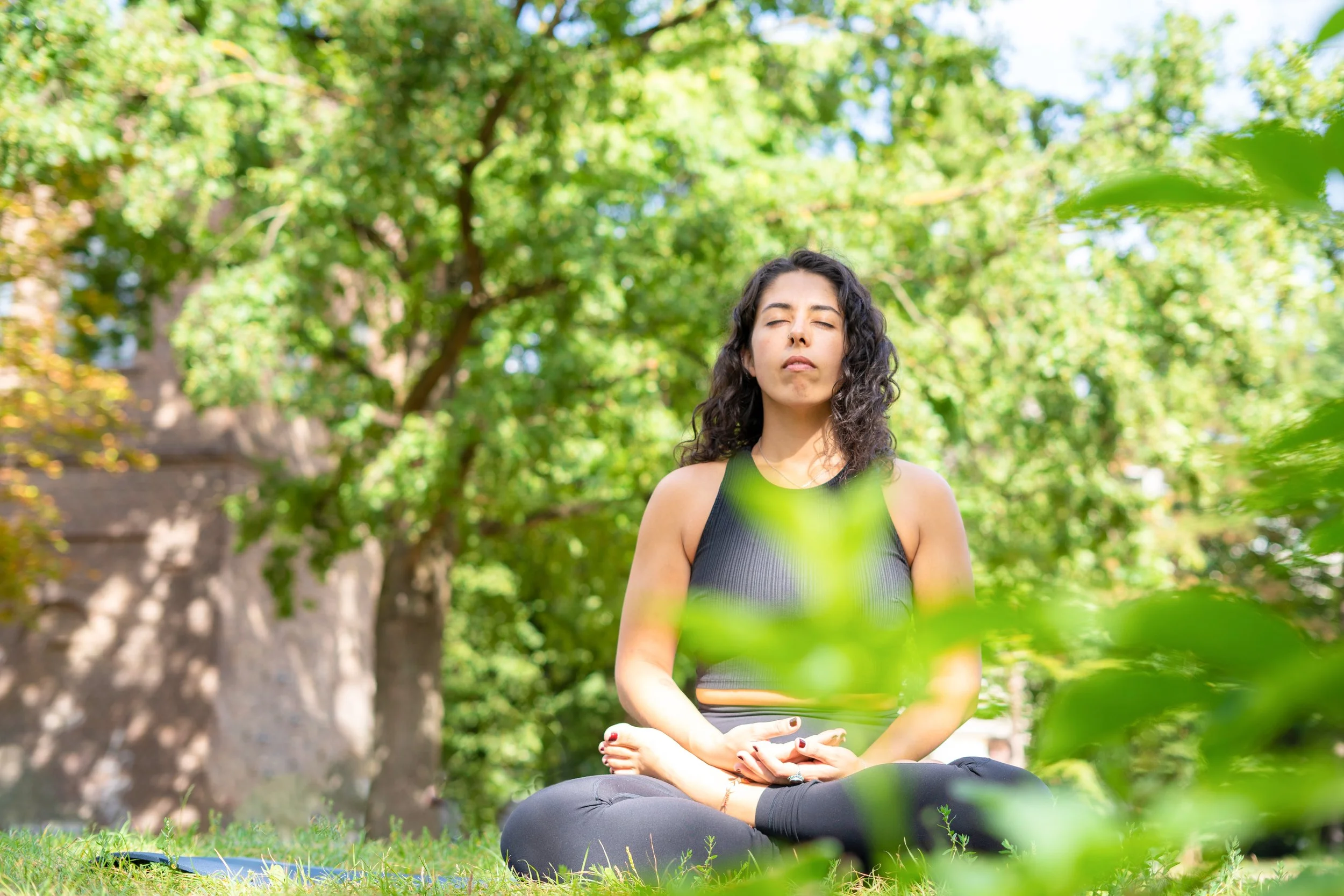 A woman practicing meditation outdoors in a park, surrounded by green trees and foliage.