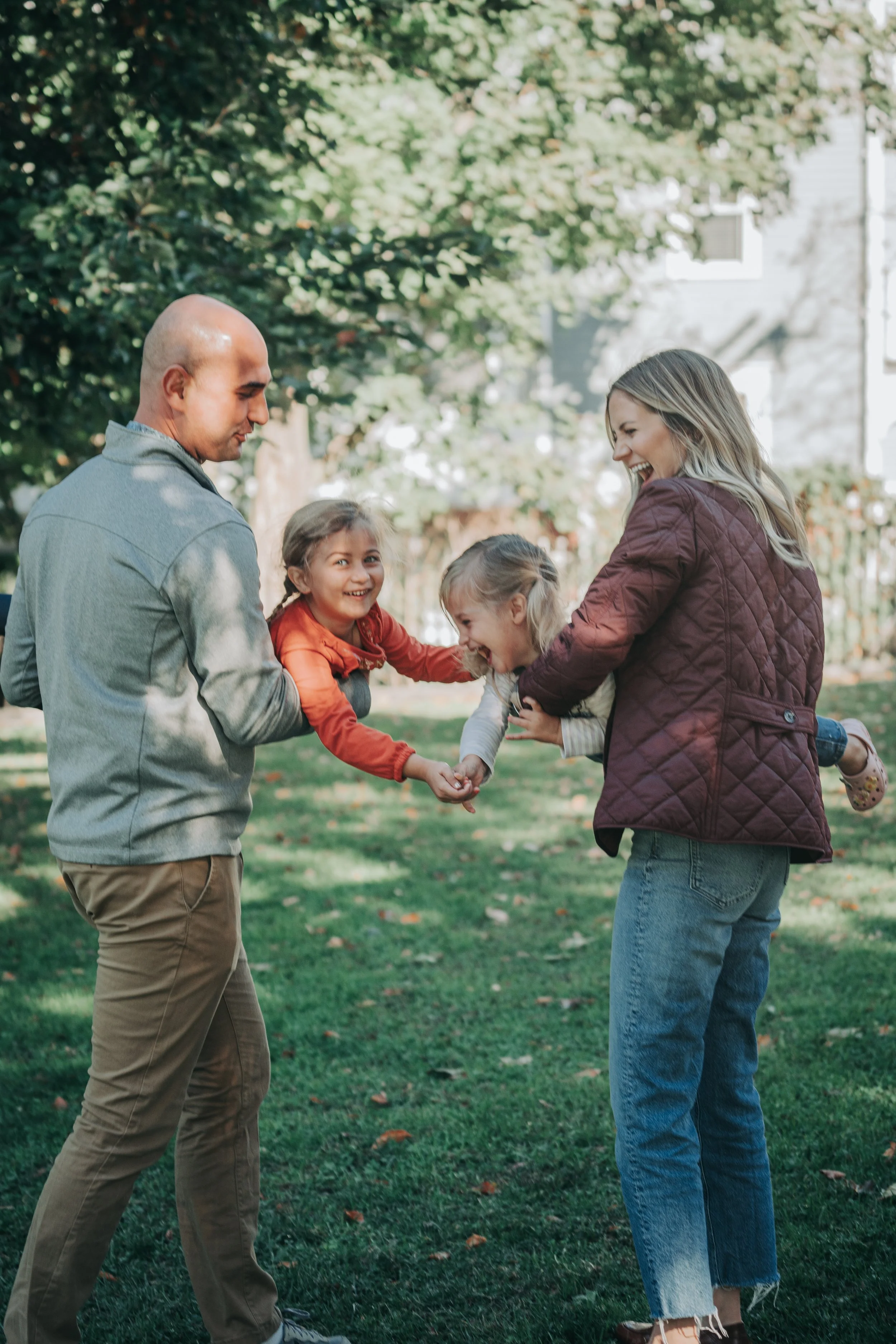 A family of four playing together outside on a sunny day, with two children and two adults smiling and holding hands, surrounded by green grass and trees.