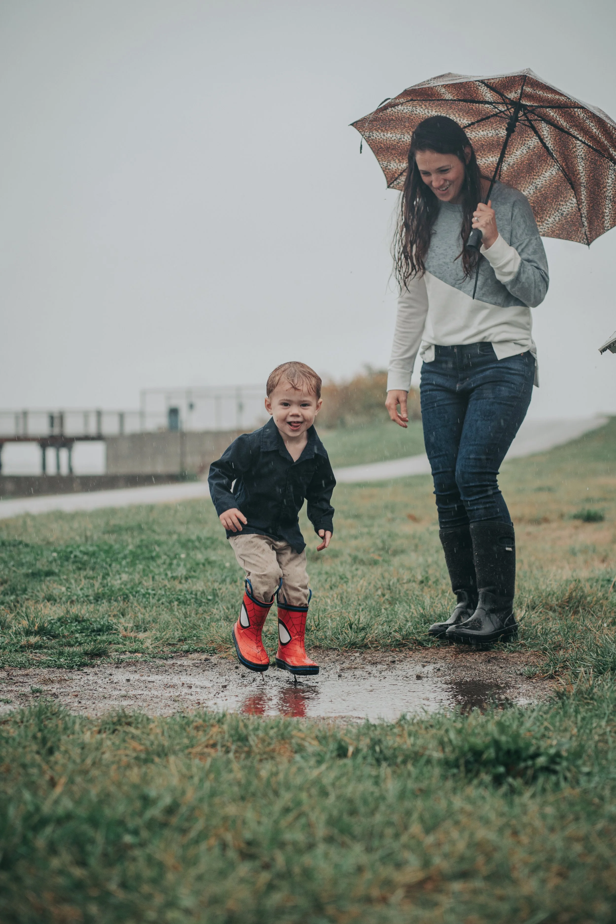 A woman and a young boy, both smiling and outdoors in rainy weather. The boy is wearing red Spiderman rain boots and is jumping over a puddle while the woman holds an umbrella, wearing rain boots.