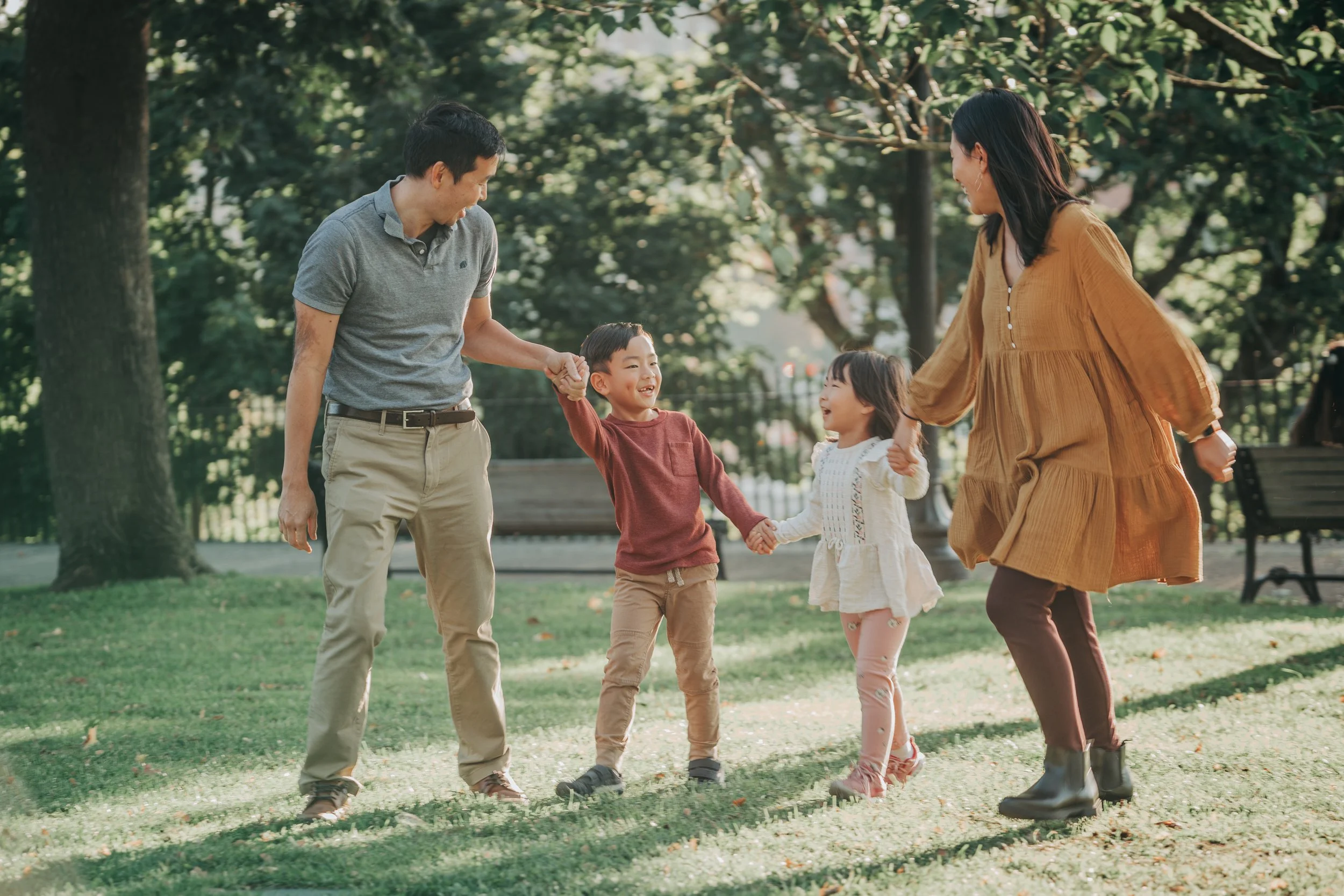 A family of four, including two children, playing and holding hands in a park with trees and benches in the background.