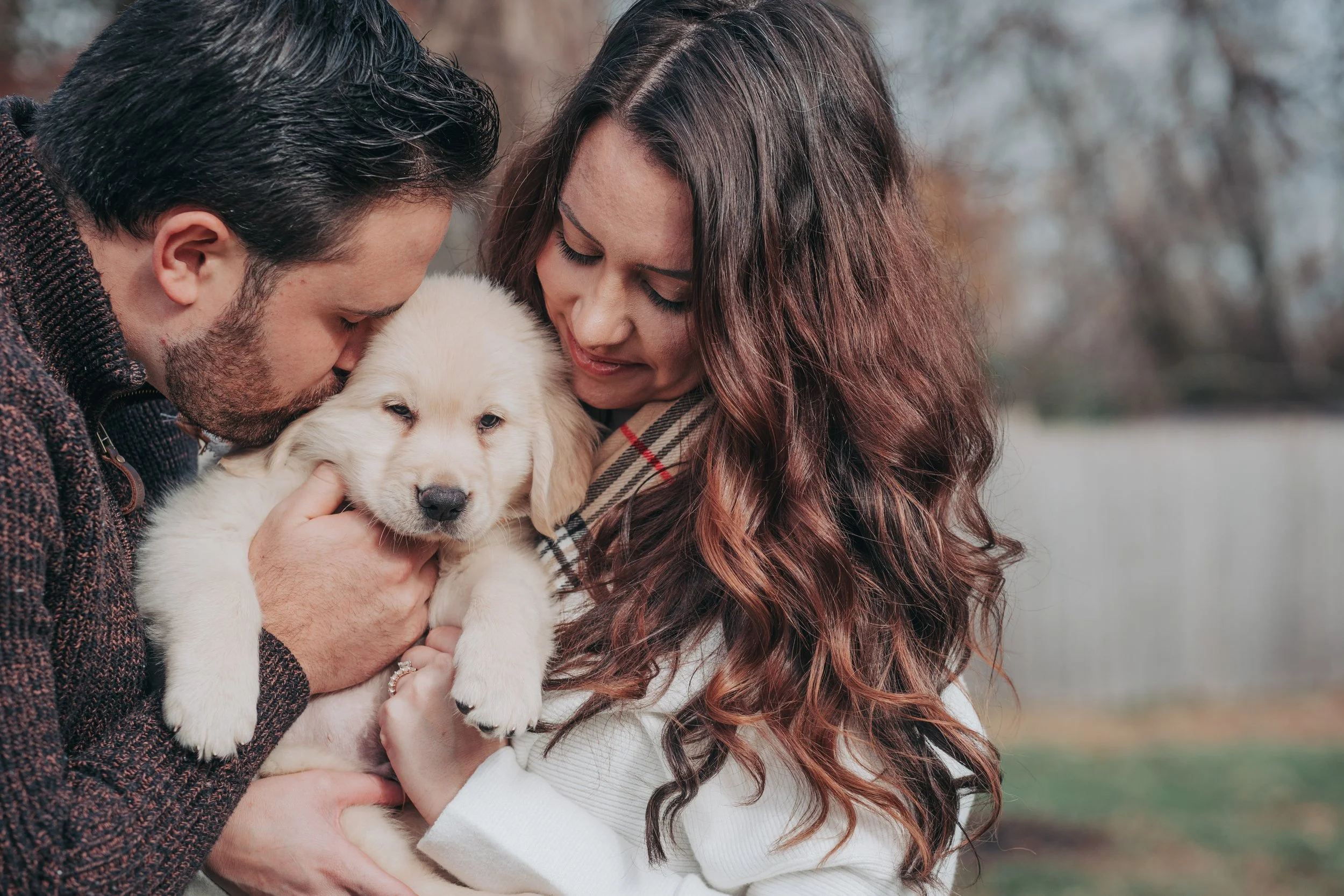 A man and woman holding a golden retriever puppy outdoors in fall with trees and a fence in the background.