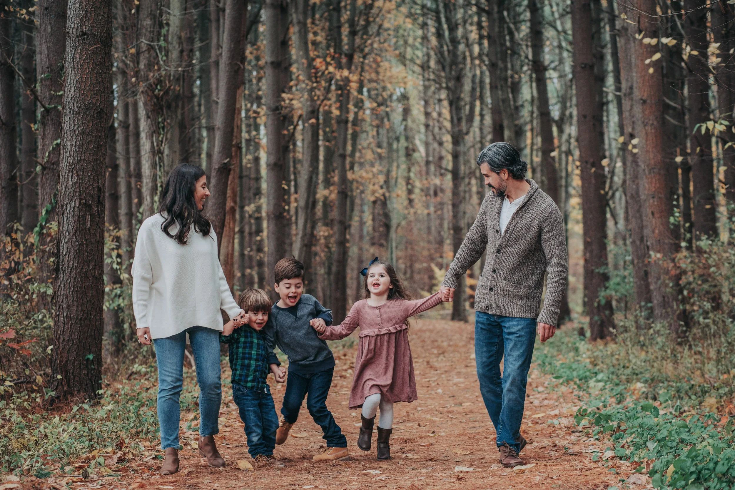 Family of five walking and playing in a forest during autumn, holding hands and smiling.