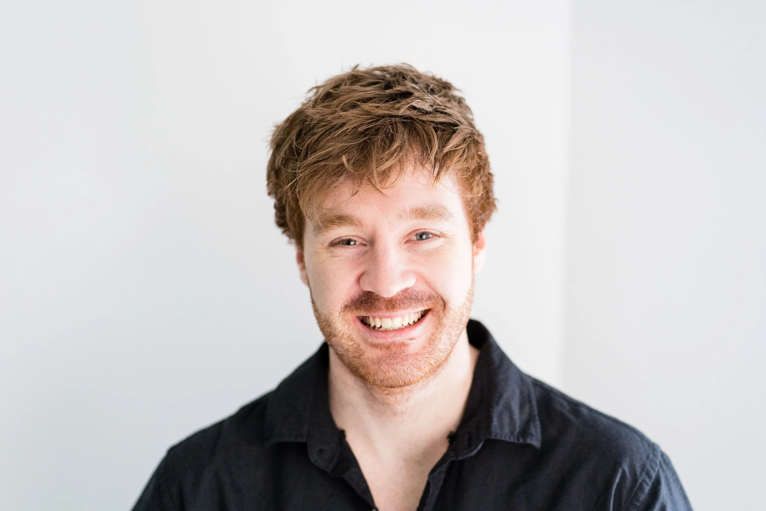 A smiling man with reddish-brown hair, a beard, and a black shirt standing against a plain white background.