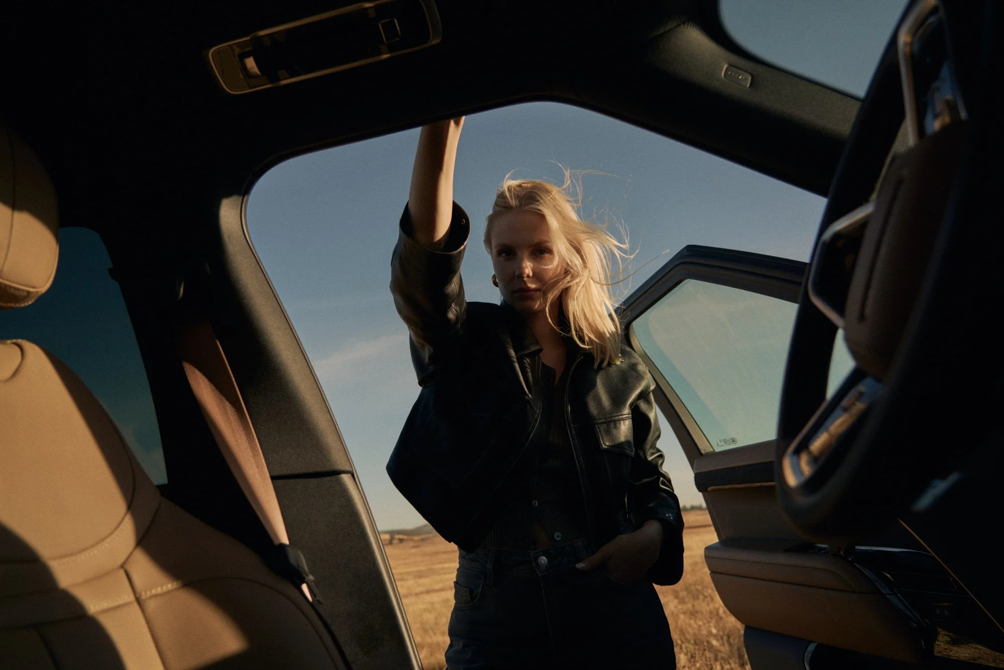 A WOMAN WITH BLONDE HAIR WEARING A BLACK LEATHER JACKET STANDING OUTSIDE A CAR WITH ITS DOOR OPEN, IN A RURAL LANDSCAPE DURING SUNSET OR SUNRISE.