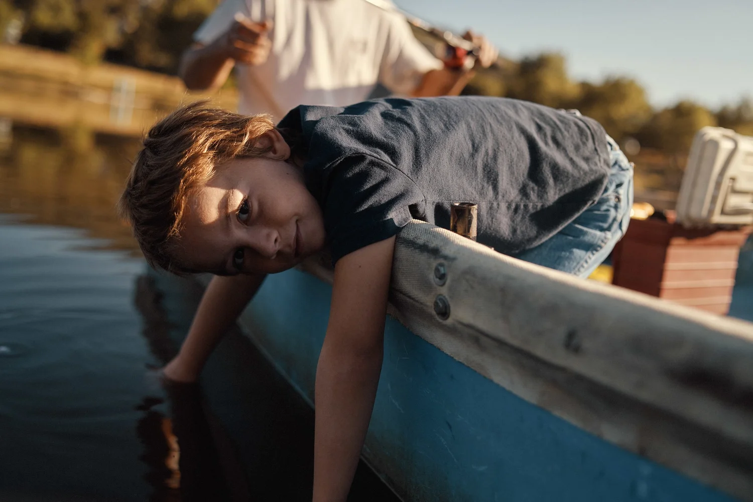 A CHILD LEANING OVER THE SIDE OF A BOAT AND LOOKING AT THE WATER, WITH AN ADULT AND TREE-LINED SHORE BEHIND.