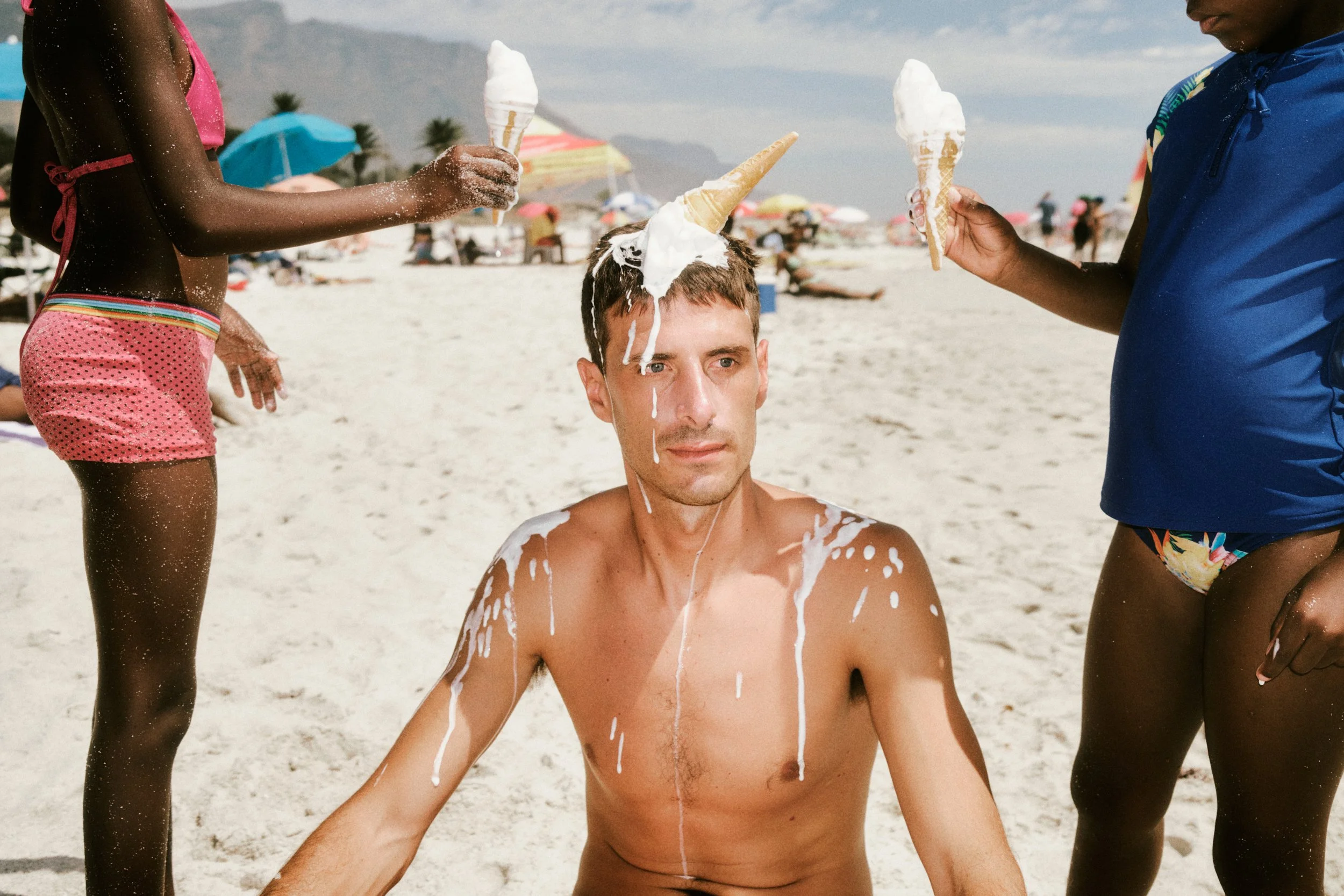 TWO CHILDREN PLACING ICE CREAM CONES ON A MAN’S HEAD ON A CAPE TOWN BEACH.