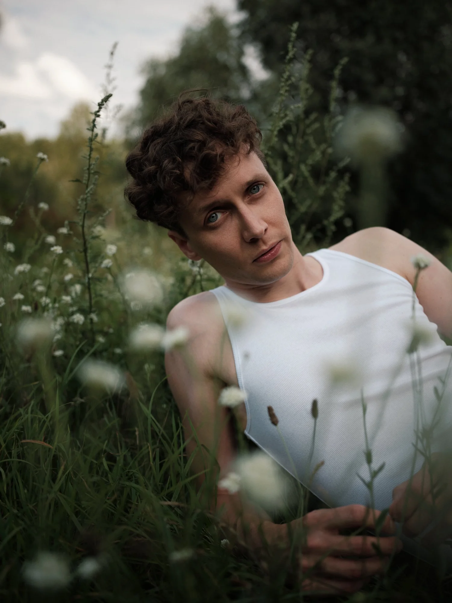 ACTOR JULIUS FELDMEIER WITH CURLY BROWN HAIR AND BLUE EYES LYING OUTDOORS IN A GRASSY FIELD SURROUNDED BY TALL PLANTS, WEARING A WHITE SLEEVELESS SHIRT.