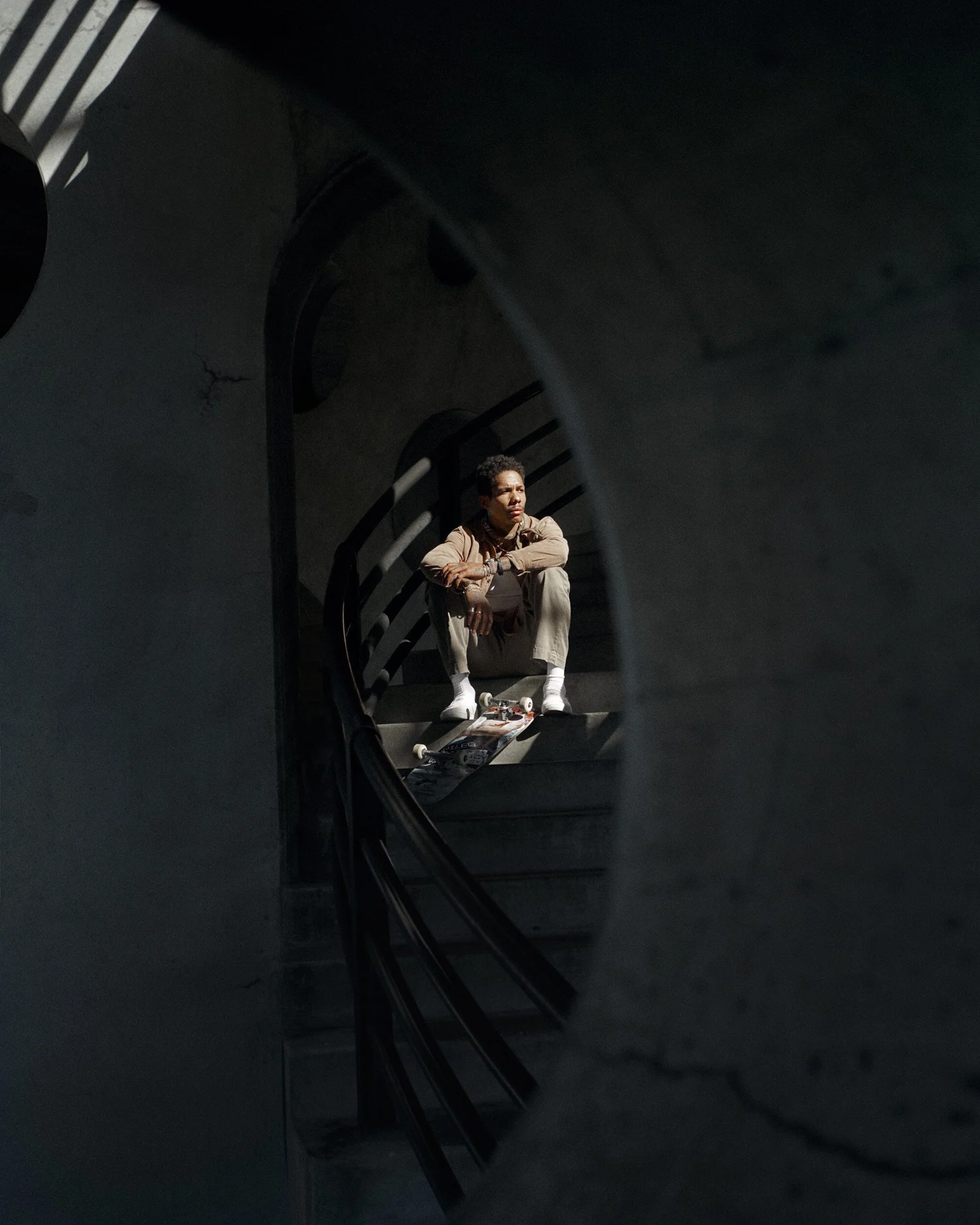 BOO JOHNSON SEATED ON STAIRS IN A TAN JACKET AND TROUSERS WITH A SKATEBOARD AT HIS FEET.