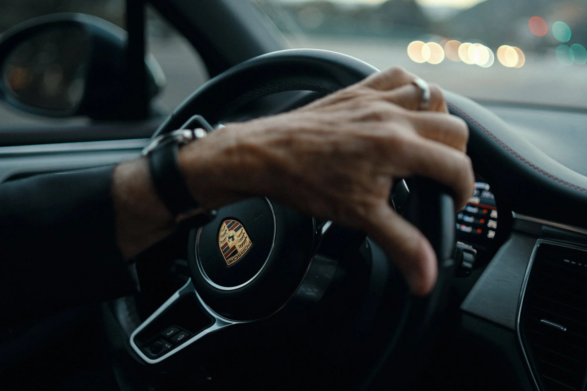 CLOSE-UP OF A PERSON'S HAND GRIPPING A PORSCHE CAR STEERING WHEEL INSIDE A VEHICLE, WITH BLURRED CITY LIGHTS VISIBLE THROUGH THE WINDSHIELD.