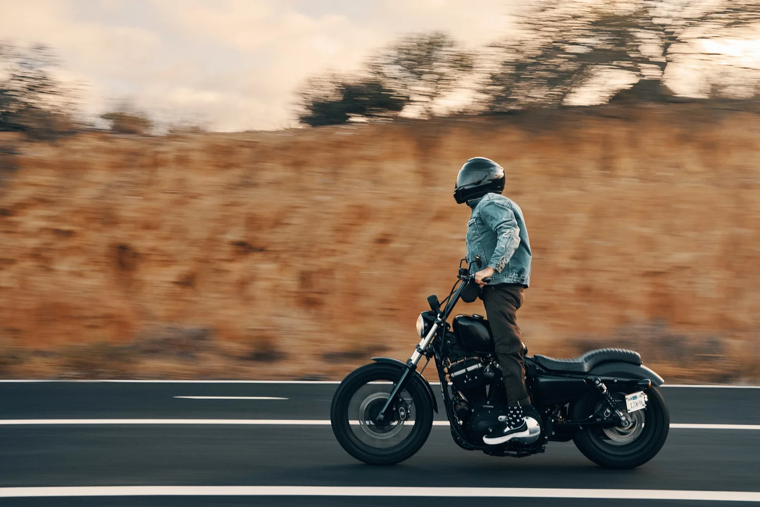 A PERSON RIDING A BLACK MOTORCYCLE ON A PAVED ROAD WITH A GRASSY HILLSIDE AND TREES IN THE BACKGROUND DURING SUNSET OR SUNRISE.